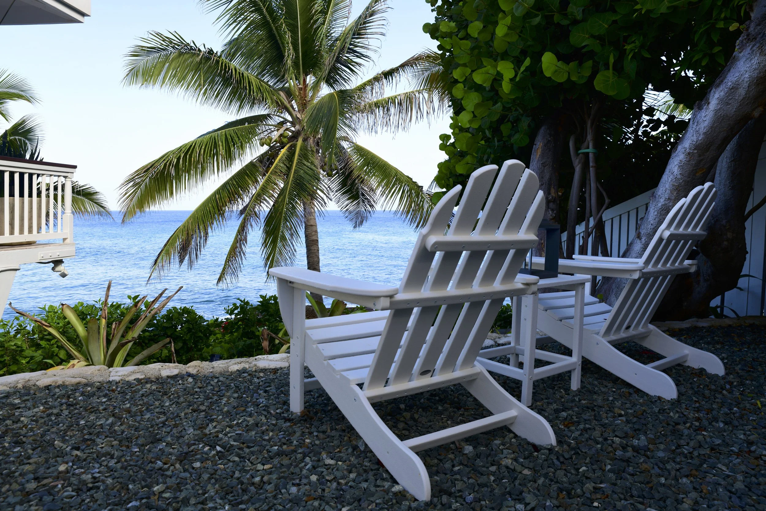 Two white Adirondack chairs on a gravel patio overlooking the ocean, with palm trees and greenery to the side.