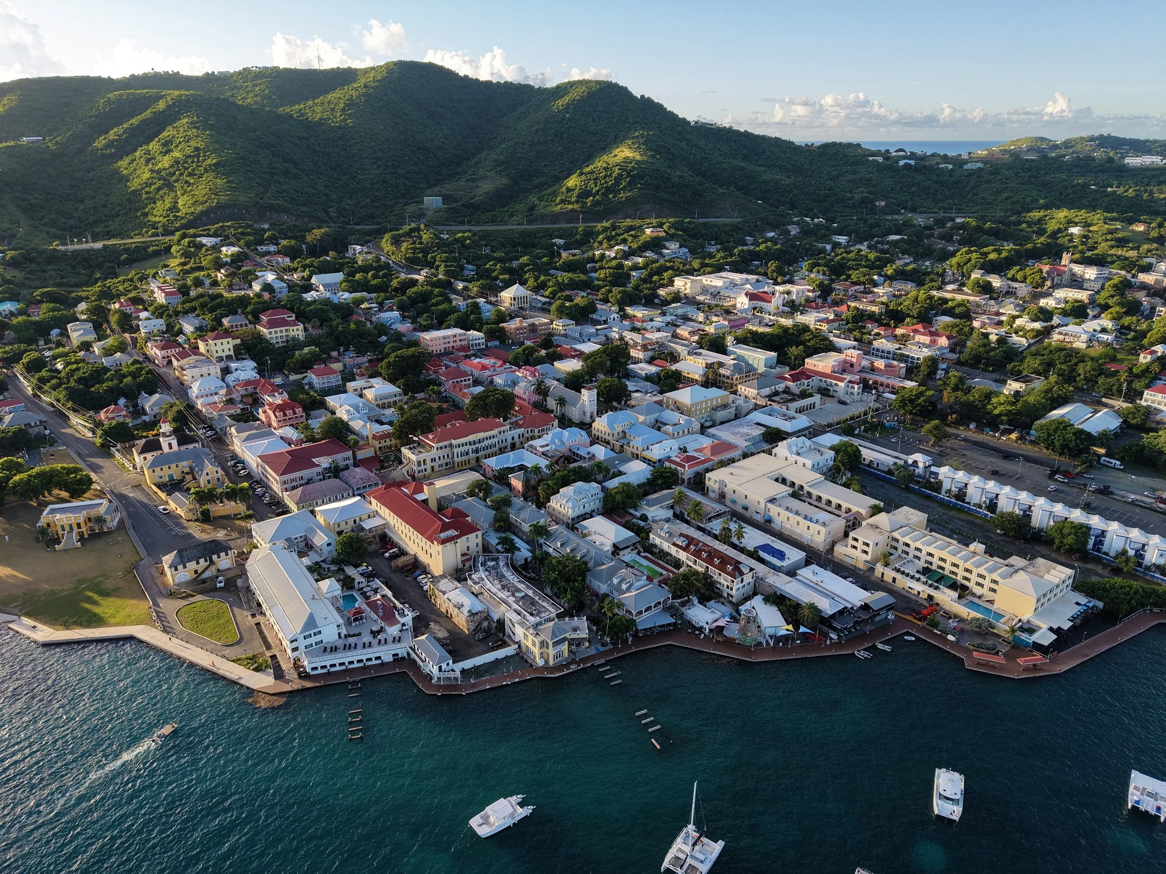 Aerial view of a coastal town with colorful buildings, boats in the water, green hills in the background, and a clear sky.