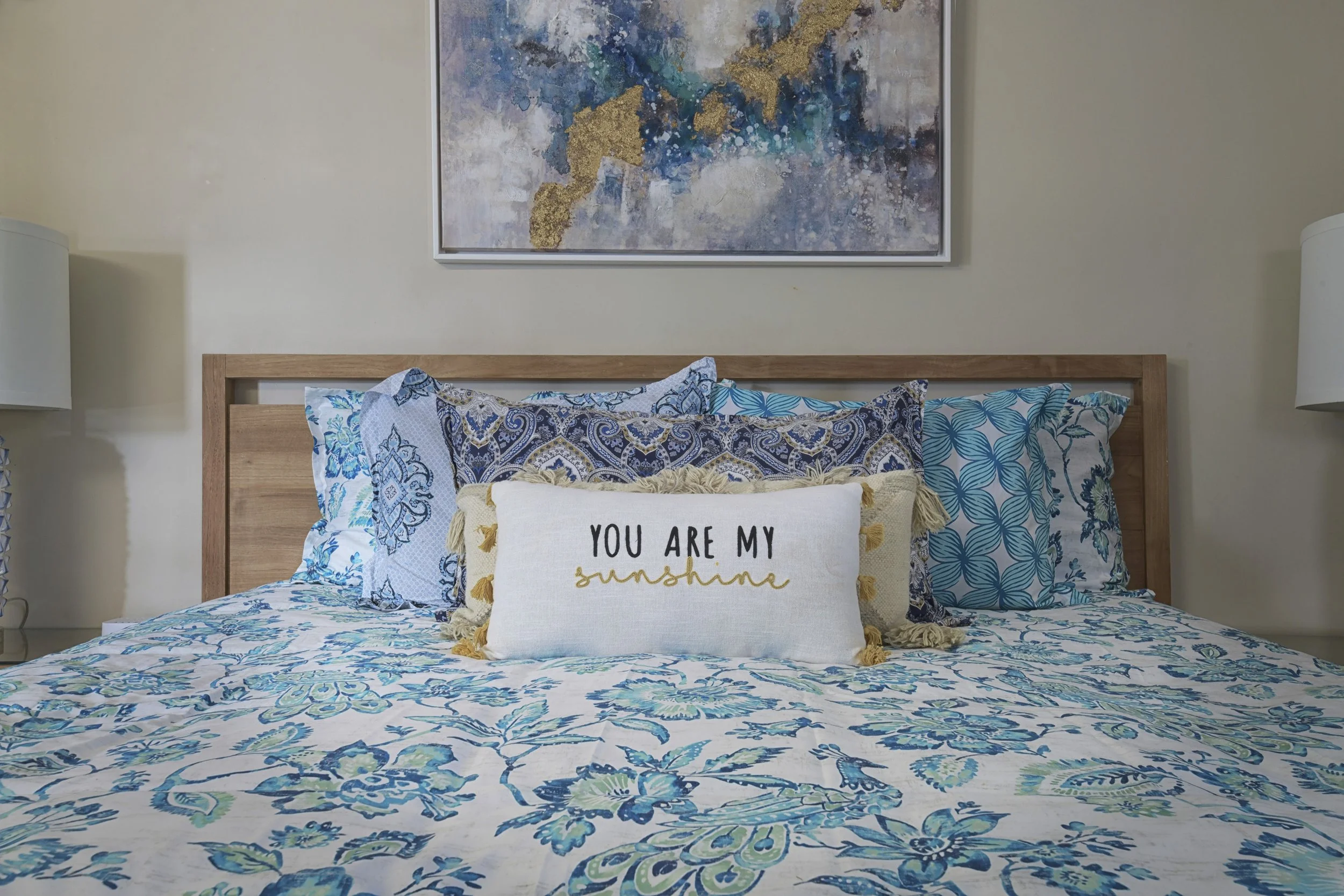 A neatly made bed with blue and white patterned bedding and multiple pillows, including a decorative pillow that reads 'You are my sunshine,' against a wall with a framed abstract painting.