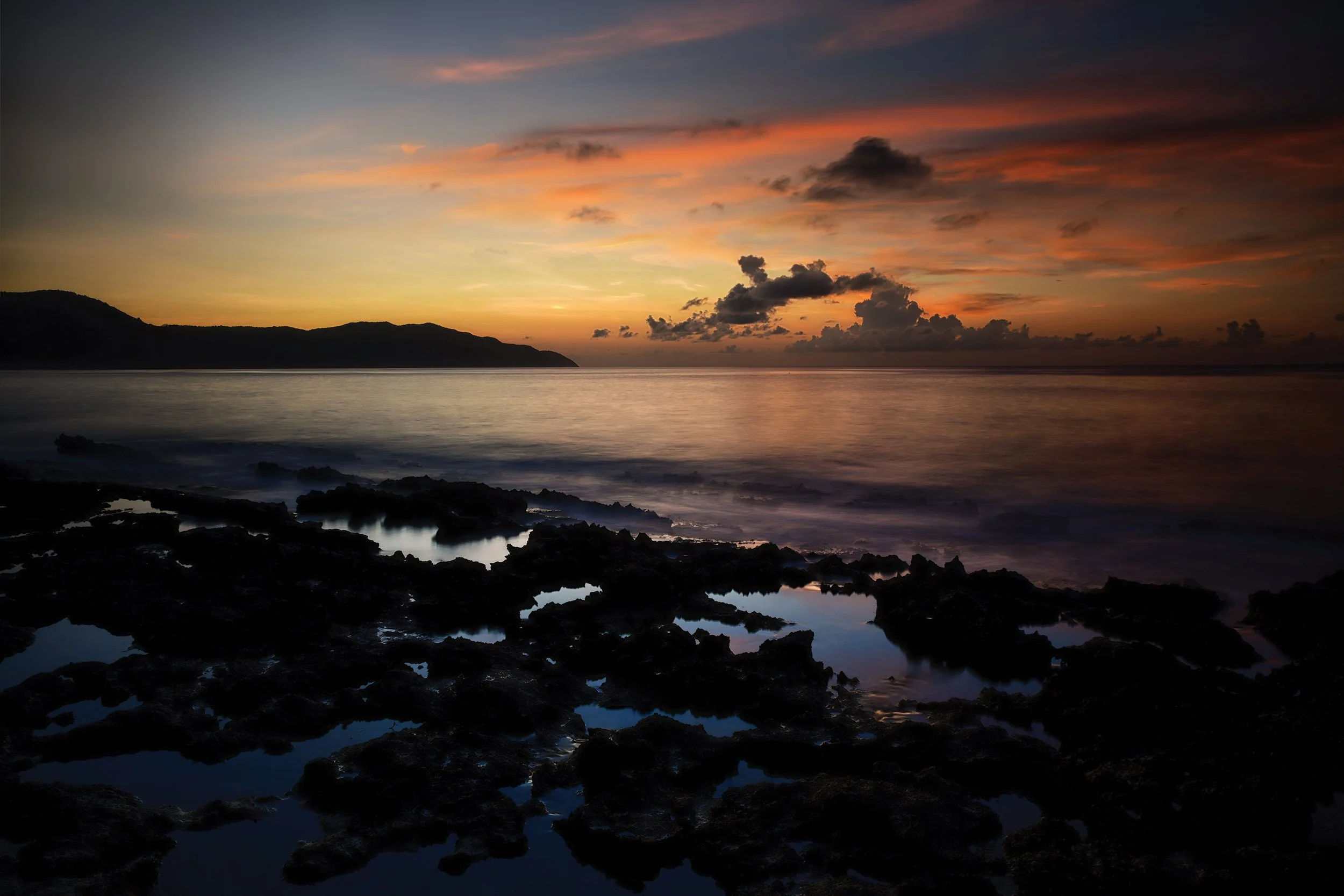 A scenic sunset over the ocean with a dark silhouette of rocky shoreline in the foreground and a mountain in the distance. The sky is filled with orange, pink, and purple hues and scattered clouds.