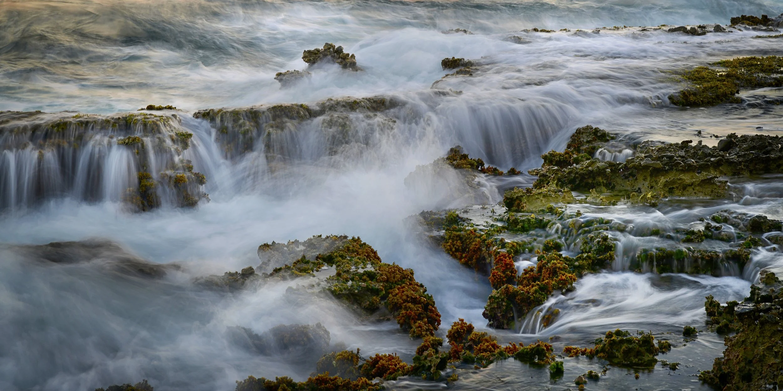 Rushing ocean waves crashing over rocks and seaweed along a shoreline.