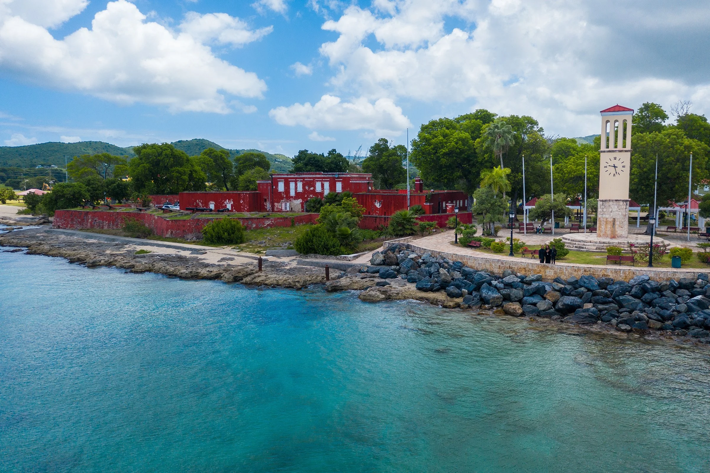 A scenic coastal view featuring a red historic building, a clock tower, and a seaside park with trees and benches, overlooking calm turquoise waters under a partly cloudy sky.