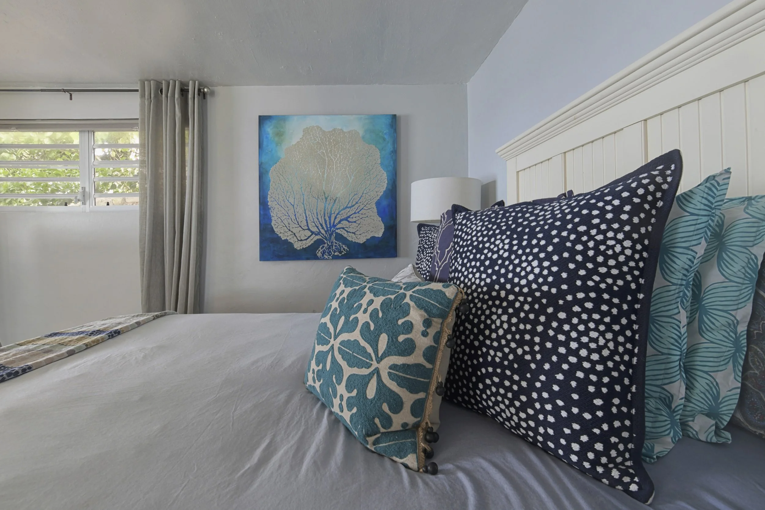 A neatly made bed with multiple decorative pillows in a bedroom with white walls, a window with louvered blinds, and a blue art piece featuring a white tree on the wall.