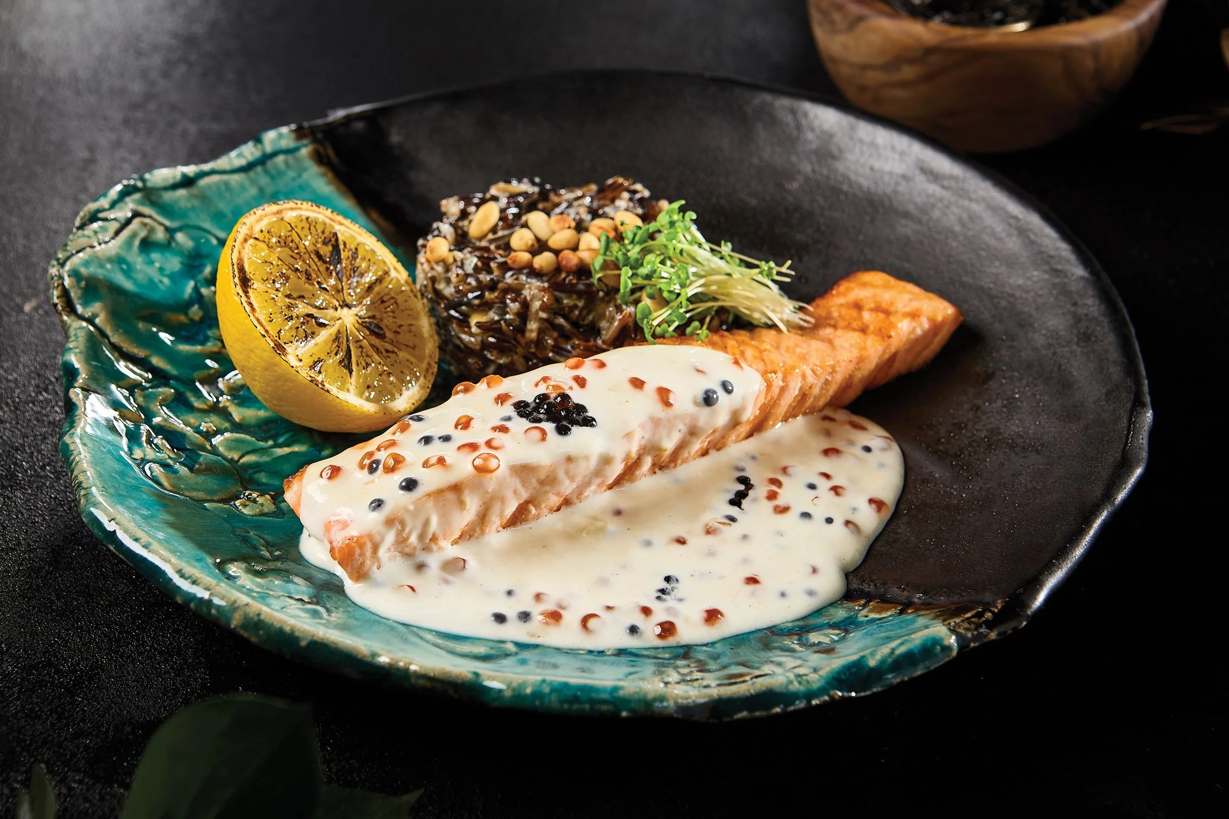 A plated dish featuring a piece of cooked salmon topped with a creamy white sauce and mustard seeds, garnished with microgreens, served with grilled lemon half, a mound of seasoned rice with pine nuts, on a black textured plate.