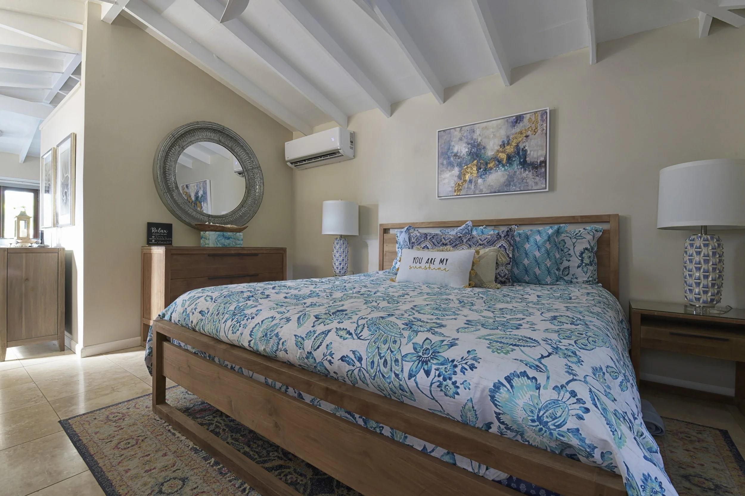 Bright bedroom with wooden bed frame, patterned blue and white bedding, decorative pillows, and a colorful abstract painting above the headboard. There are nightstands with lamps on each side, a large round mirror, and a window letting in natural lig