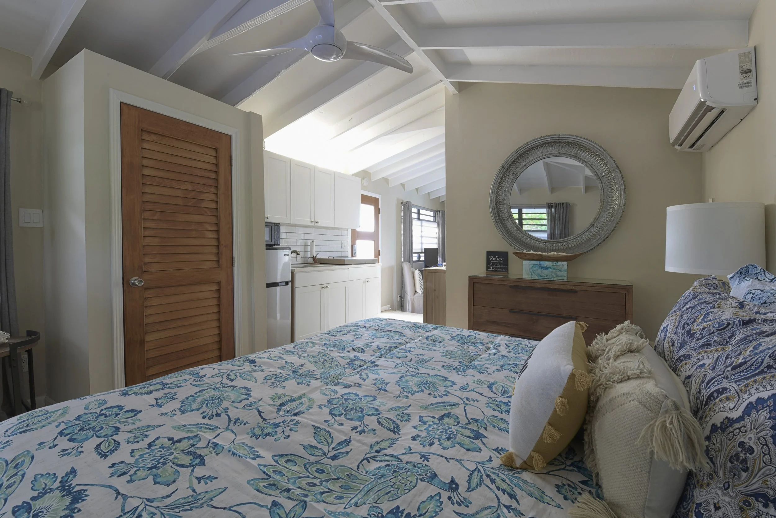 A cozy bedroom with a bed that has a blue floral quilt and decorative pillows. There's a circular mirror on the wall above a wooden dresser. The room has a sloped ceiling with white beams, a ceiling fan, and an air conditioning unit on the wall. The 