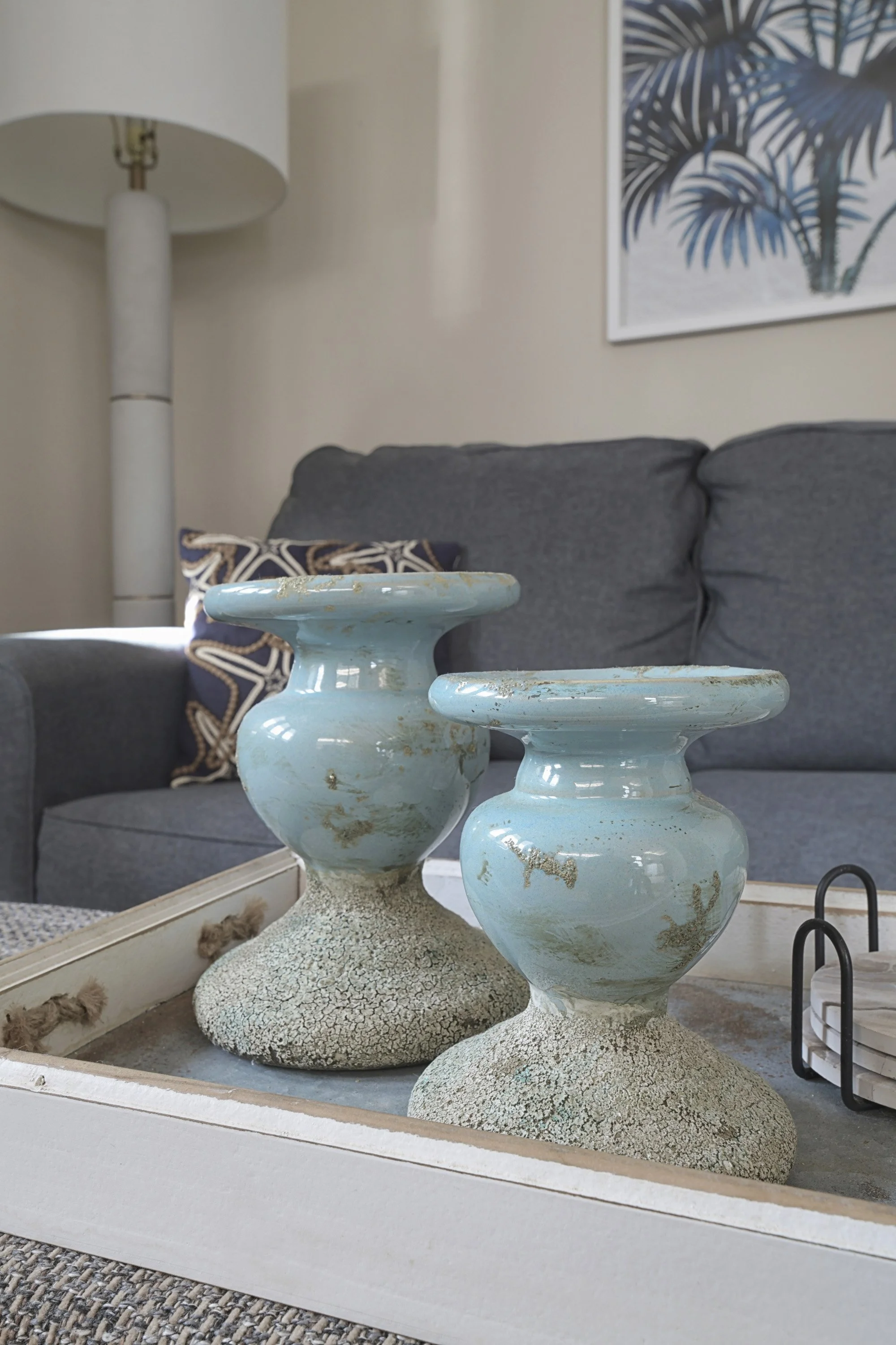 Decorative light blue ceramic vases with textured bases on a white tray on a coffee table in a living room with a gray sofa, patterned throw pillow, a lamp, and a palm print artwork on the wall.