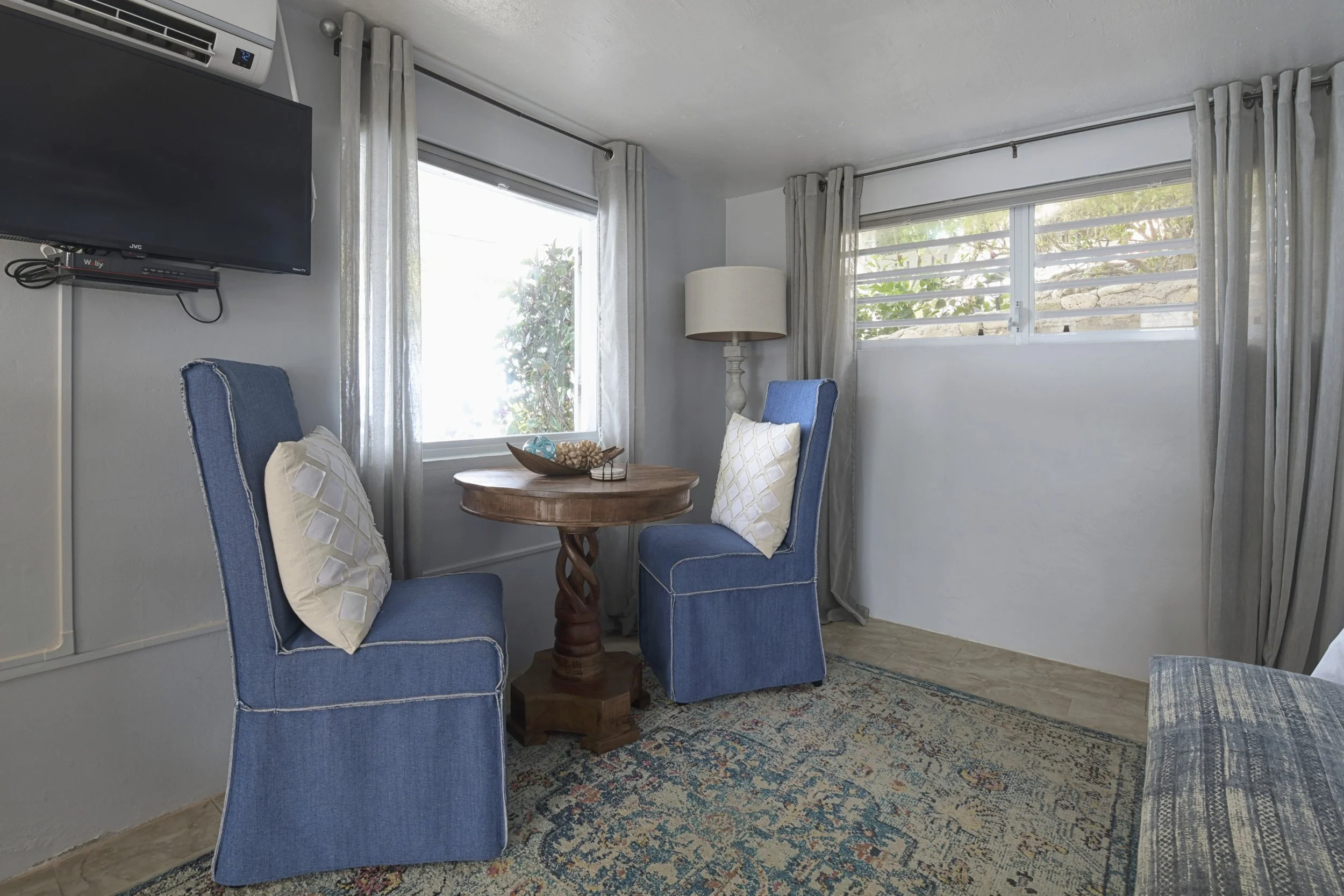A small sitting area with two blue upholstered chairs with white pillows, a round wooden table with decorative items, a floor lamp, and windows with light-colored curtains.