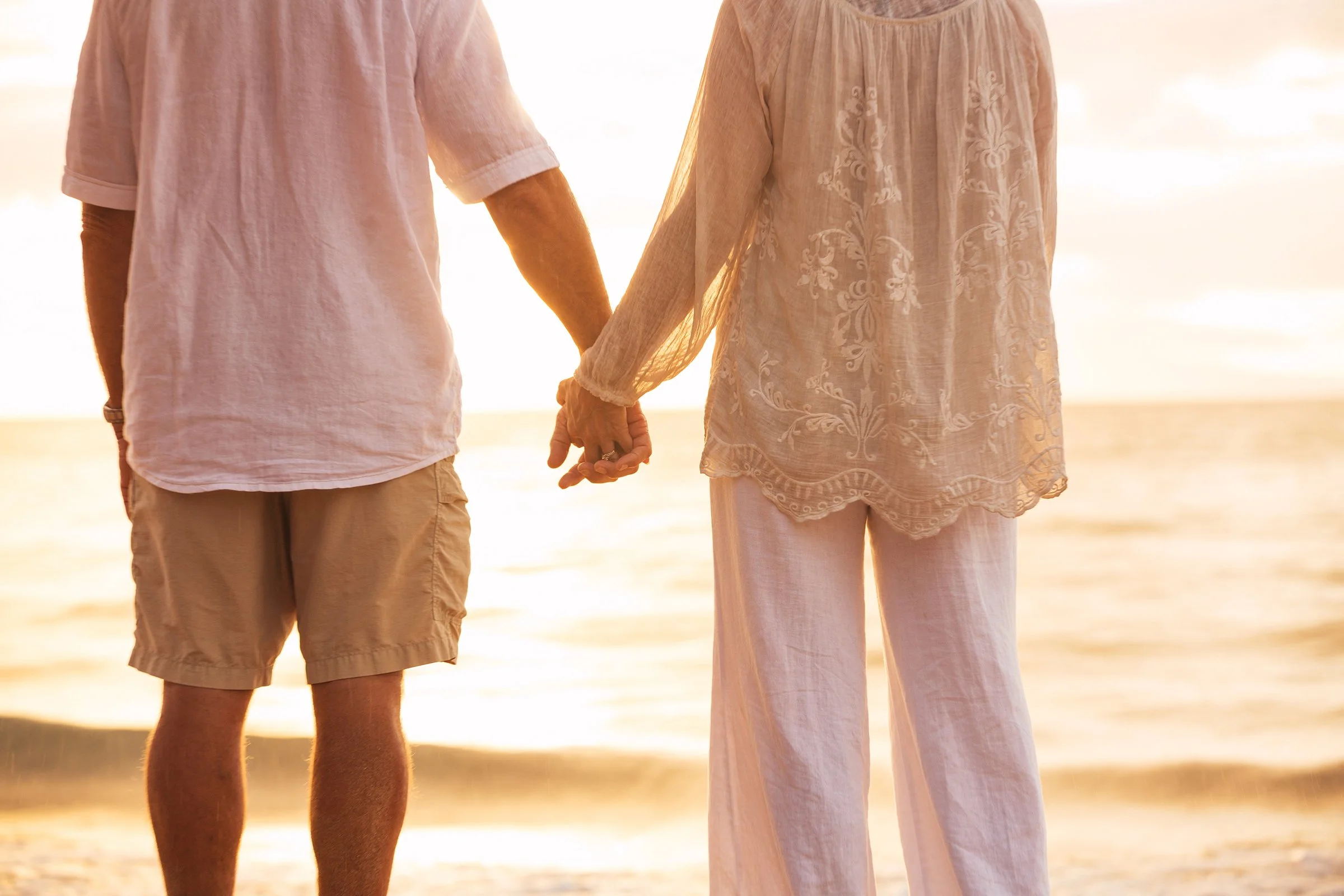 A couple holding hands on the beach during sunset, both dressed in light, summery clothing.