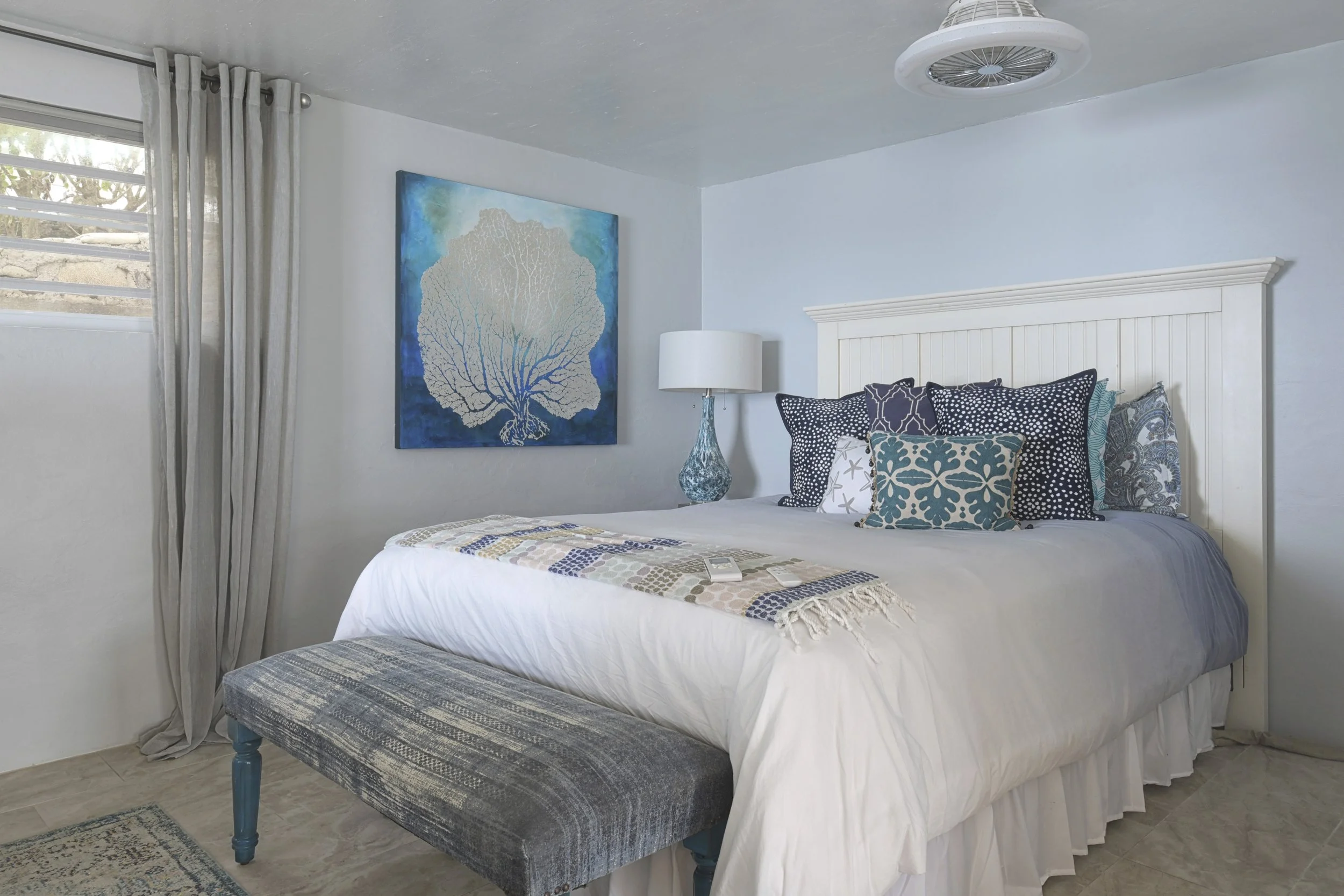 A bedroom with a white wooden headboard, decorated with multiple patterned pillows, a white bedspread, and a quilt at the foot. There is a blue and white abstract painting of a tree on the wall, a blue glass lamp on the bedside table, a window with l