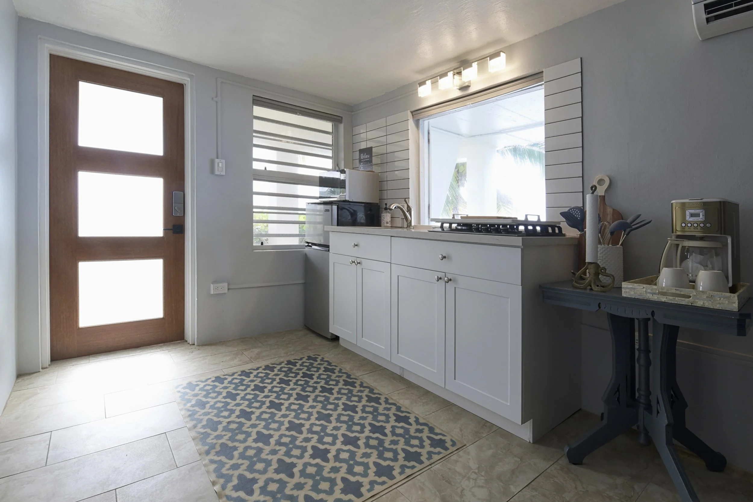 Modern kitchen with white cabinets, a small countertop, and a window with blinds, near a wooden door with four frosted glass panels, a patterned rug on tan tiled floor, and a black side table with a coffee station.