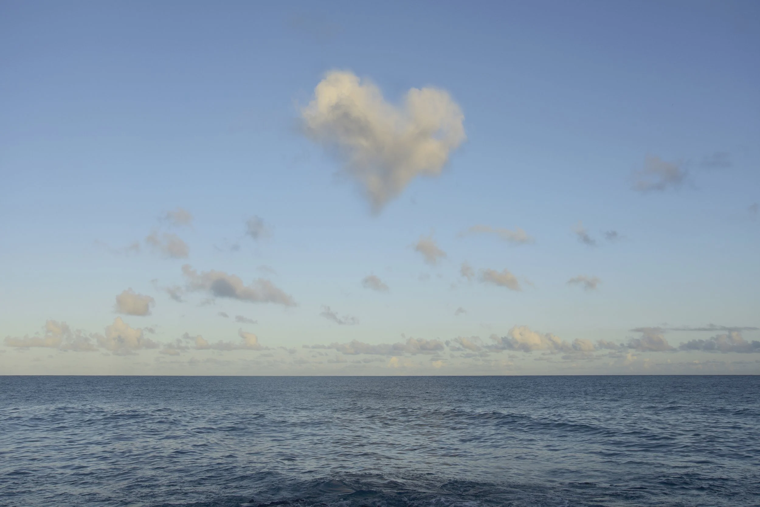 A view of the ocean with gentle waves, a blue sky with scattered clouds, and a heart-shaped cloud forming in the sky.