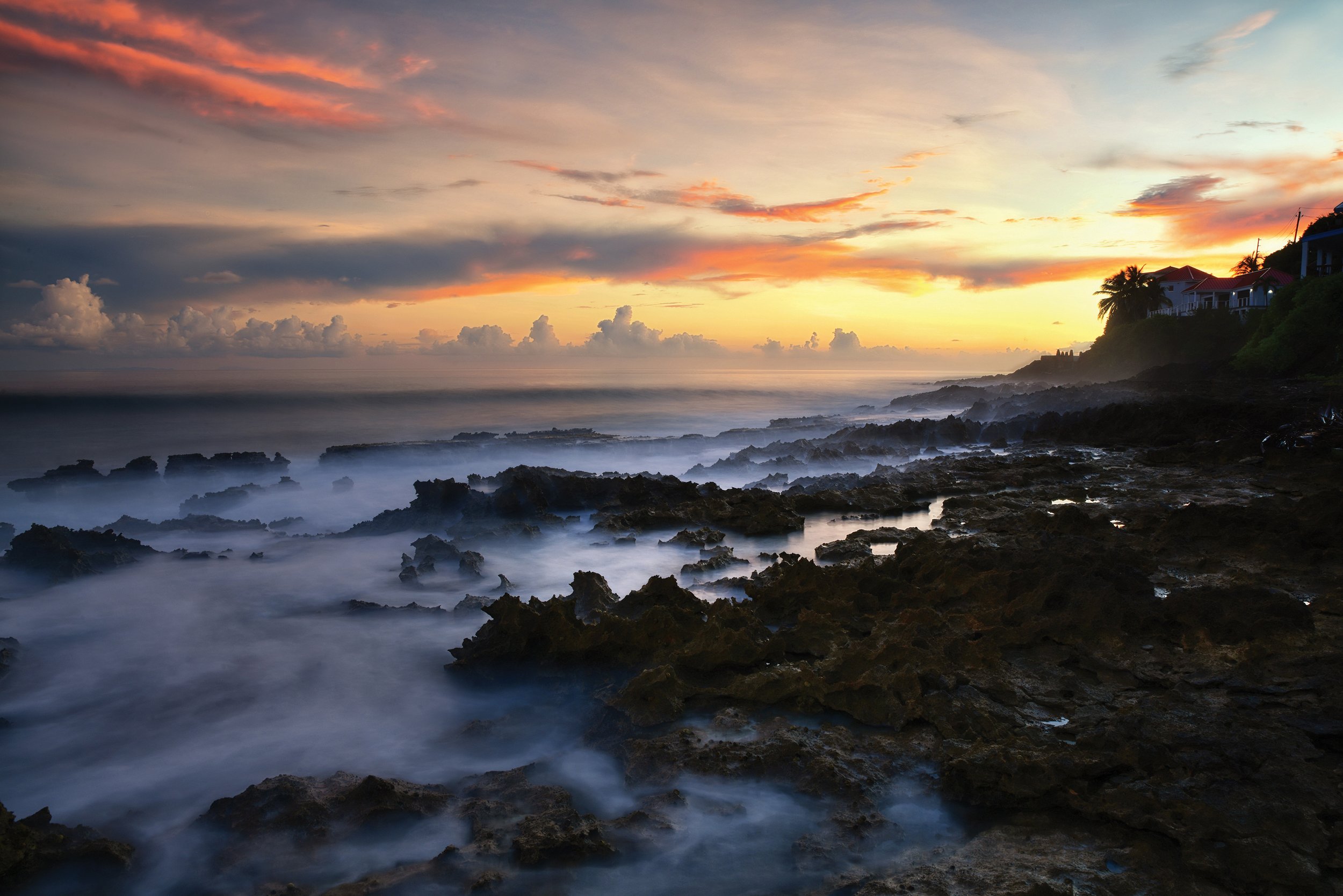 A coastal scene at sunset with rocky shoreline, misty ocean waves, and houses with palm trees on a hillside.