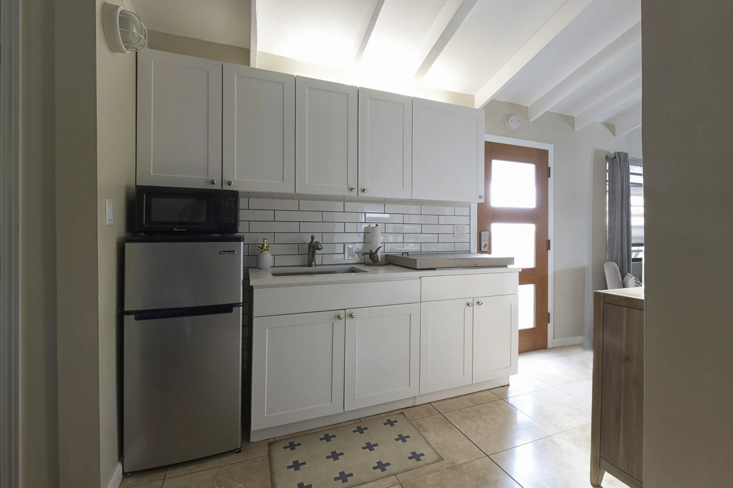 A small kitchen with white cabinets, a stainless steel refrigerator, a black microwave on top, and a white subway tile backsplash. There is a wooden door and a window with gray curtains, with a beige tiled floor and a small rug.