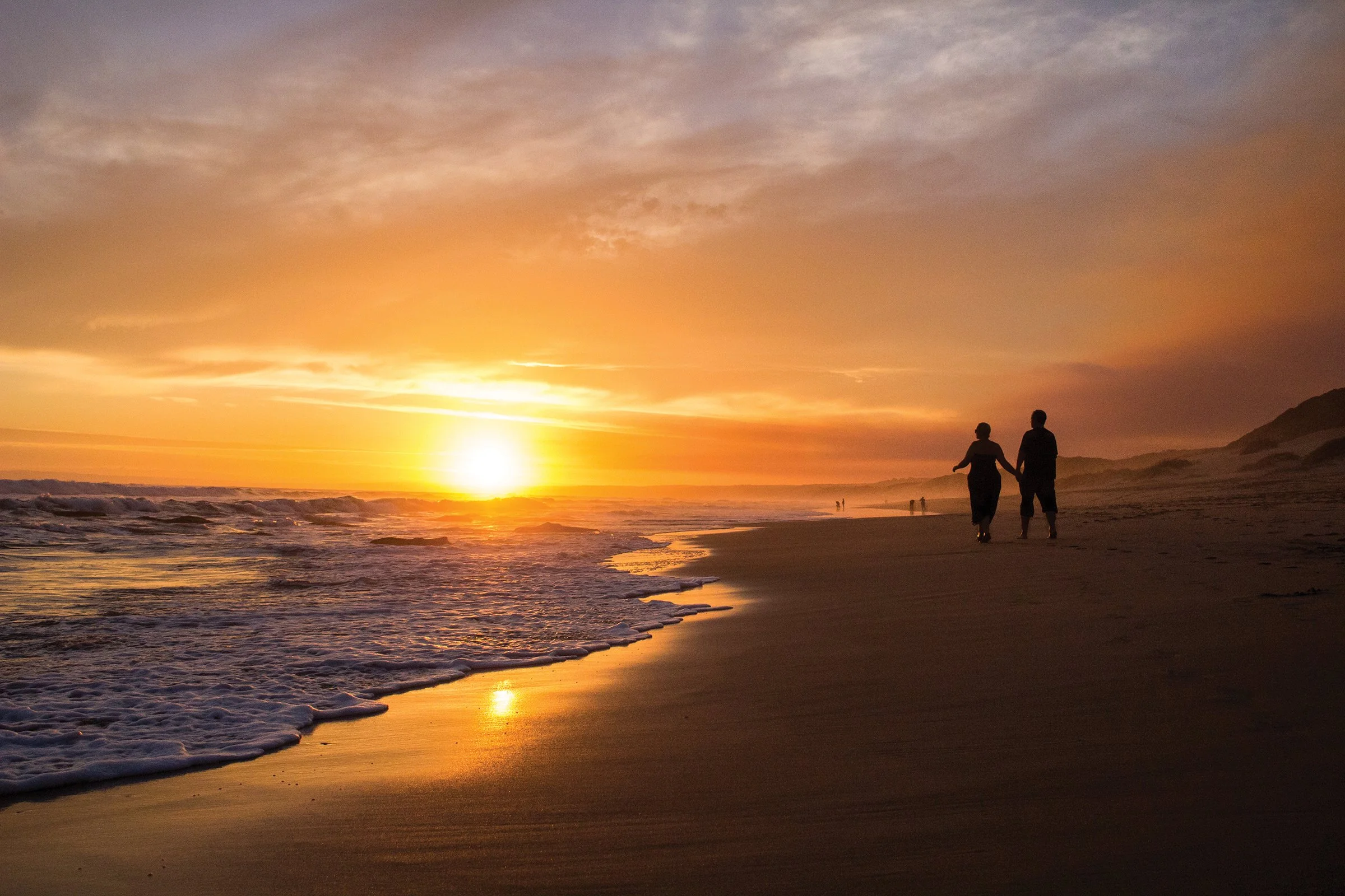 Couple walking hand in hand along a sandy beach during sunset with a colorful sky and distant shoreline.