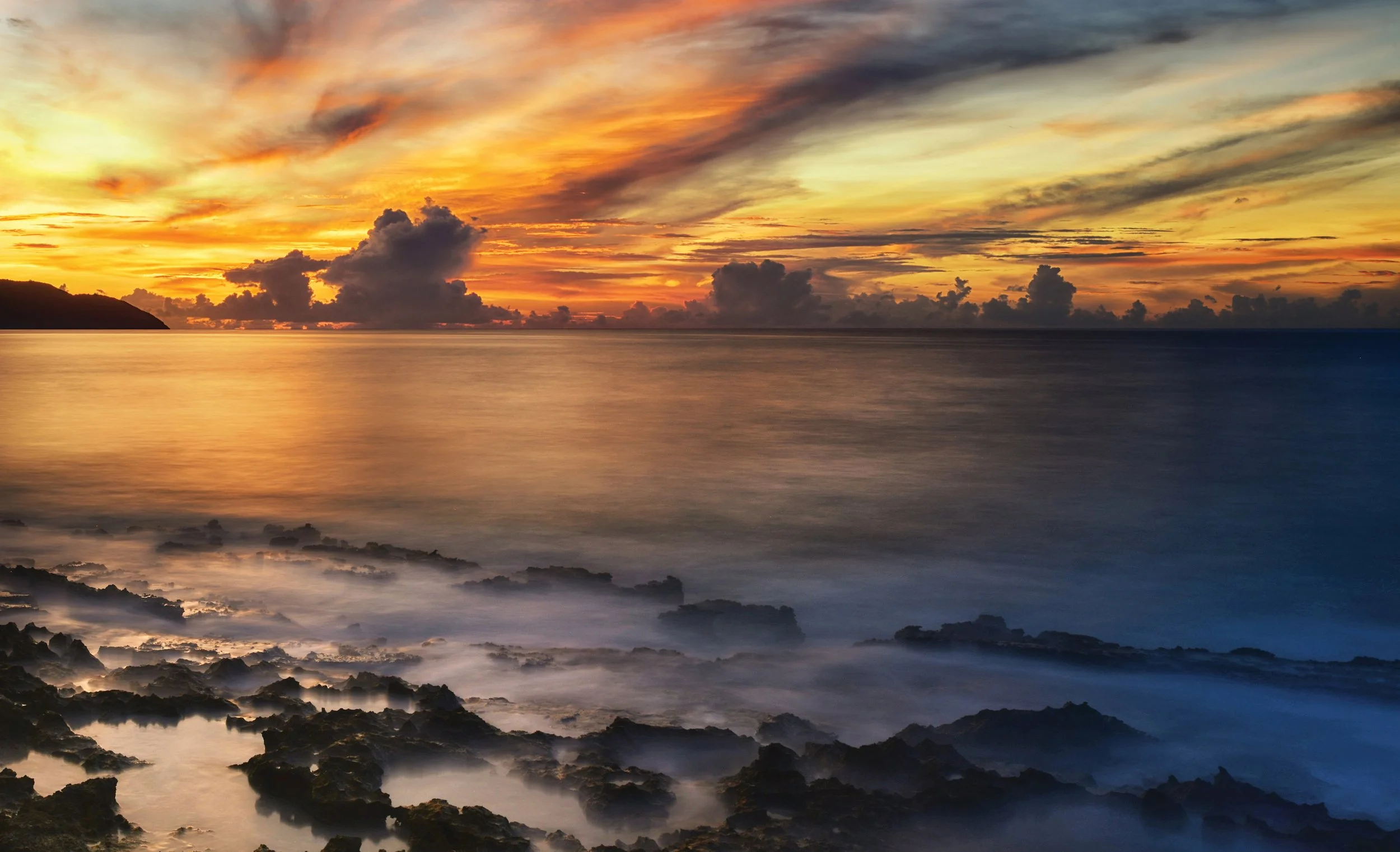Sunset over the ocean with clouds, rocky shoreline in the foreground, and a colorful sky with orange, yellow, and blue hues.