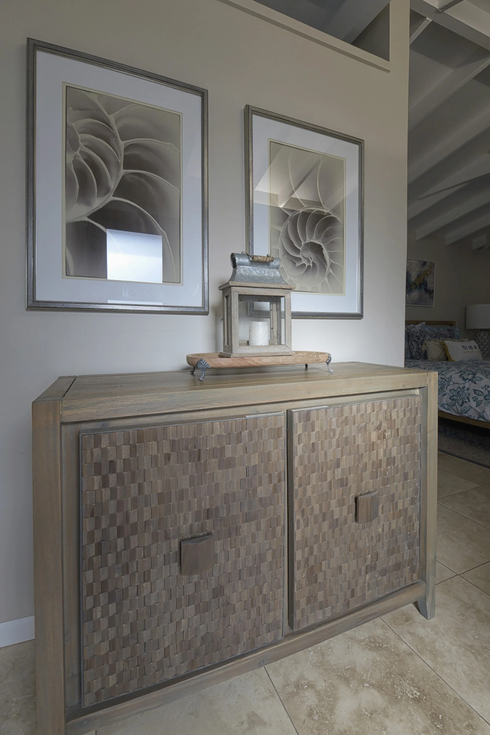 A wooden cabinet with two textured doors, topped with a decorative lantern and a wooden tray, framed abstract artwork of spiral shells on the wall, with part of a bedroom visible in the background.