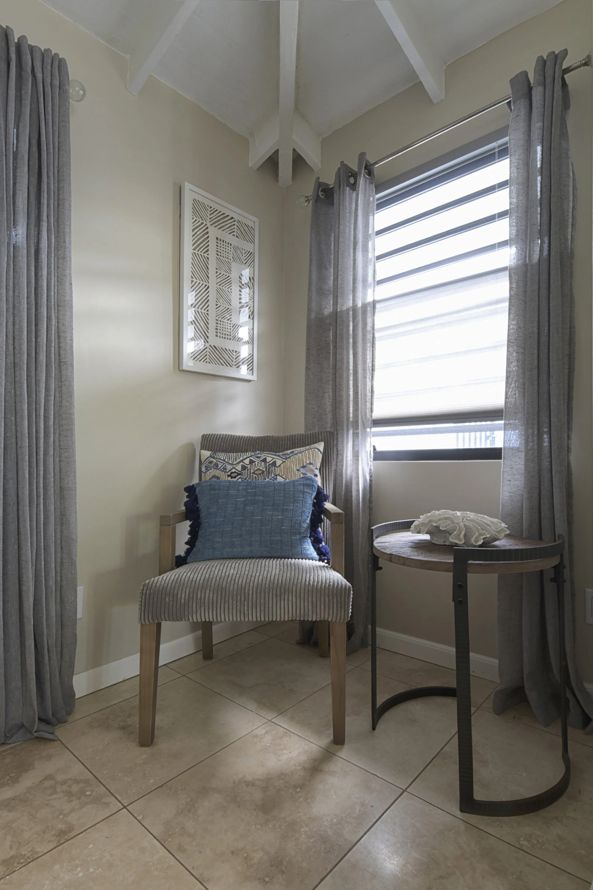 A cozy corner with beige walls and large tiled floor, featuring a gray upholstered chair with striped fabric and decorative pillows, a small round wooden table with a coral ornament, and window with sheer gray curtains and blinds, sunlight streaming 