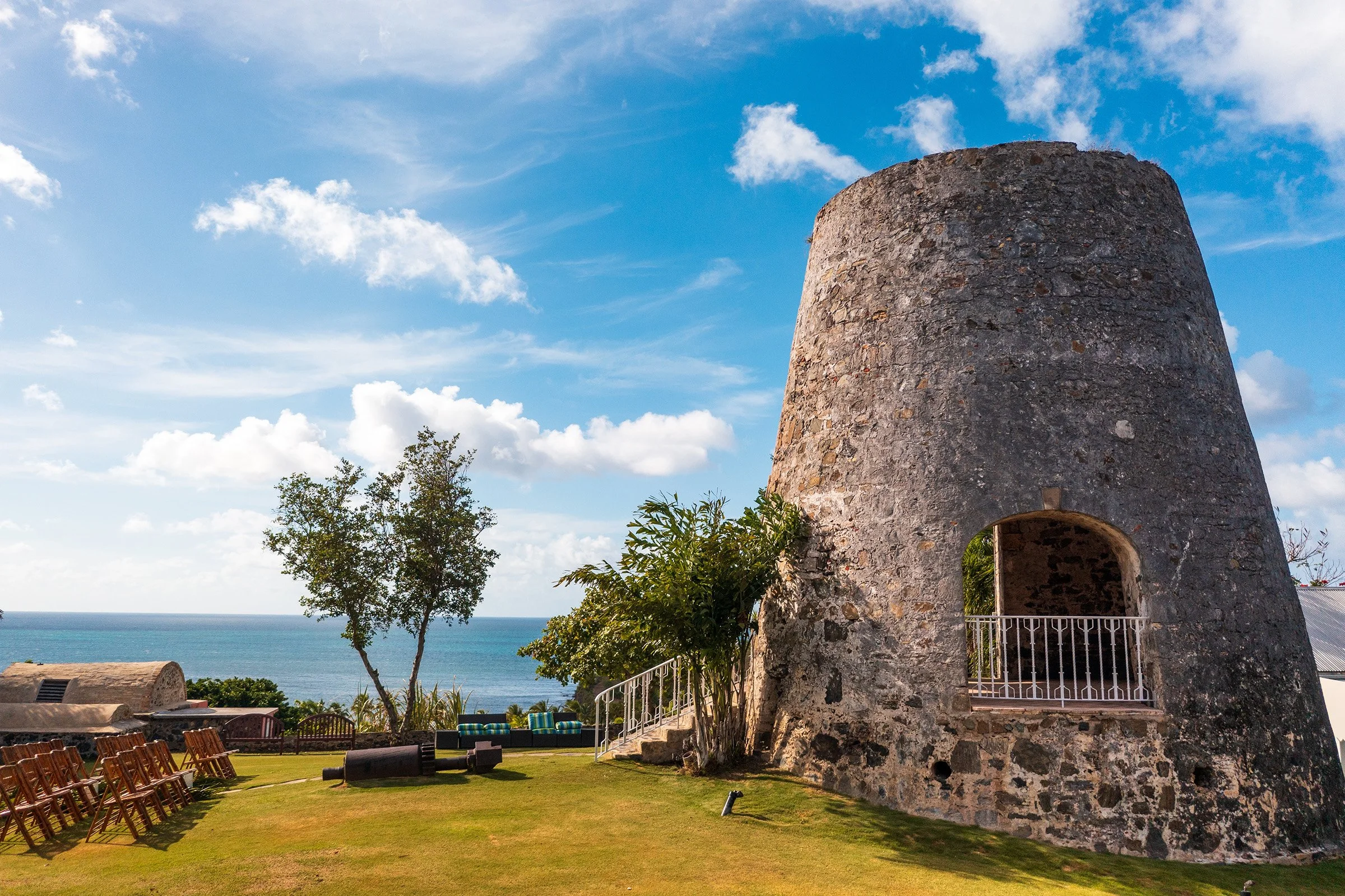 A historic stone tower overlooking the ocean, surrounded by green grass, trees, and outdoor seating, with a partly cloudy sky in the background.