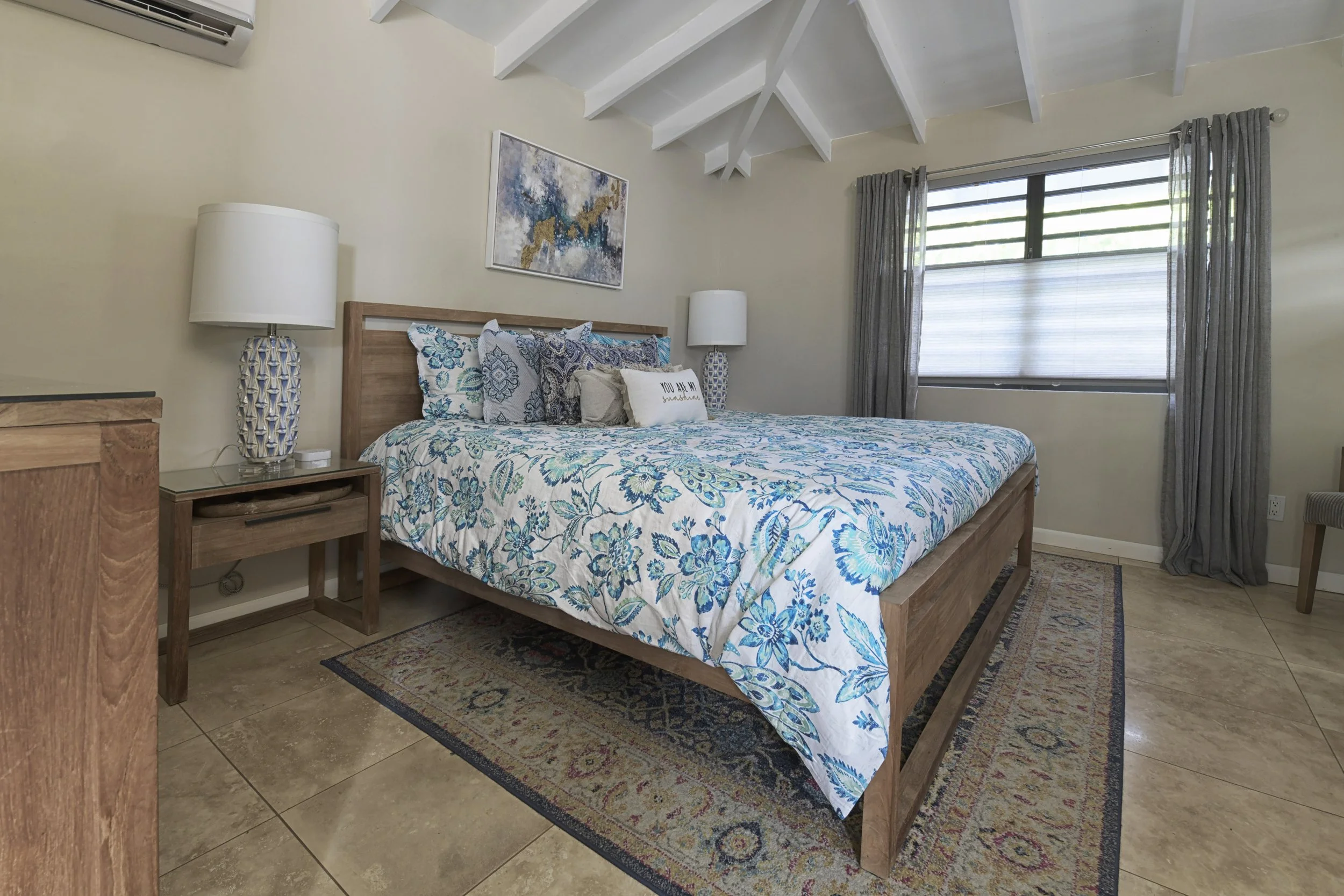 A well-made bed with a blue and white floral bedspread in a bedroom with beige walls and a high vaulted ceiling with white beams. Two matching nightstands with lamps are on either side of the bed, with a window and gray curtains on the right side of 