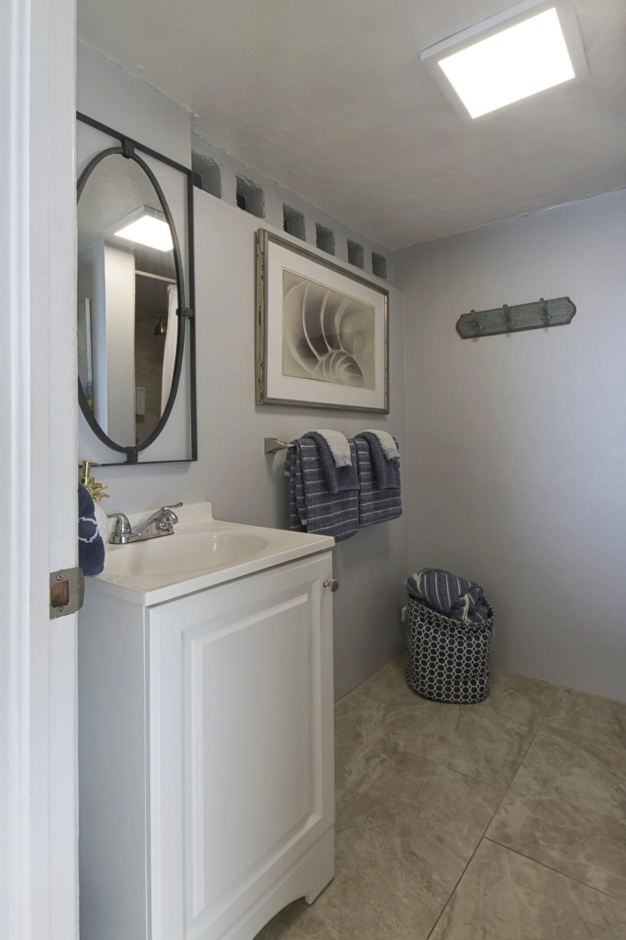 A small bathroom with a white vanity, black framed mirror, and towel rack with blue and white towels. There is framed artwork on the wall, a decorative wall piece, and a laundry basket with blue towels inside. The floor has beige tiles and the ceilin