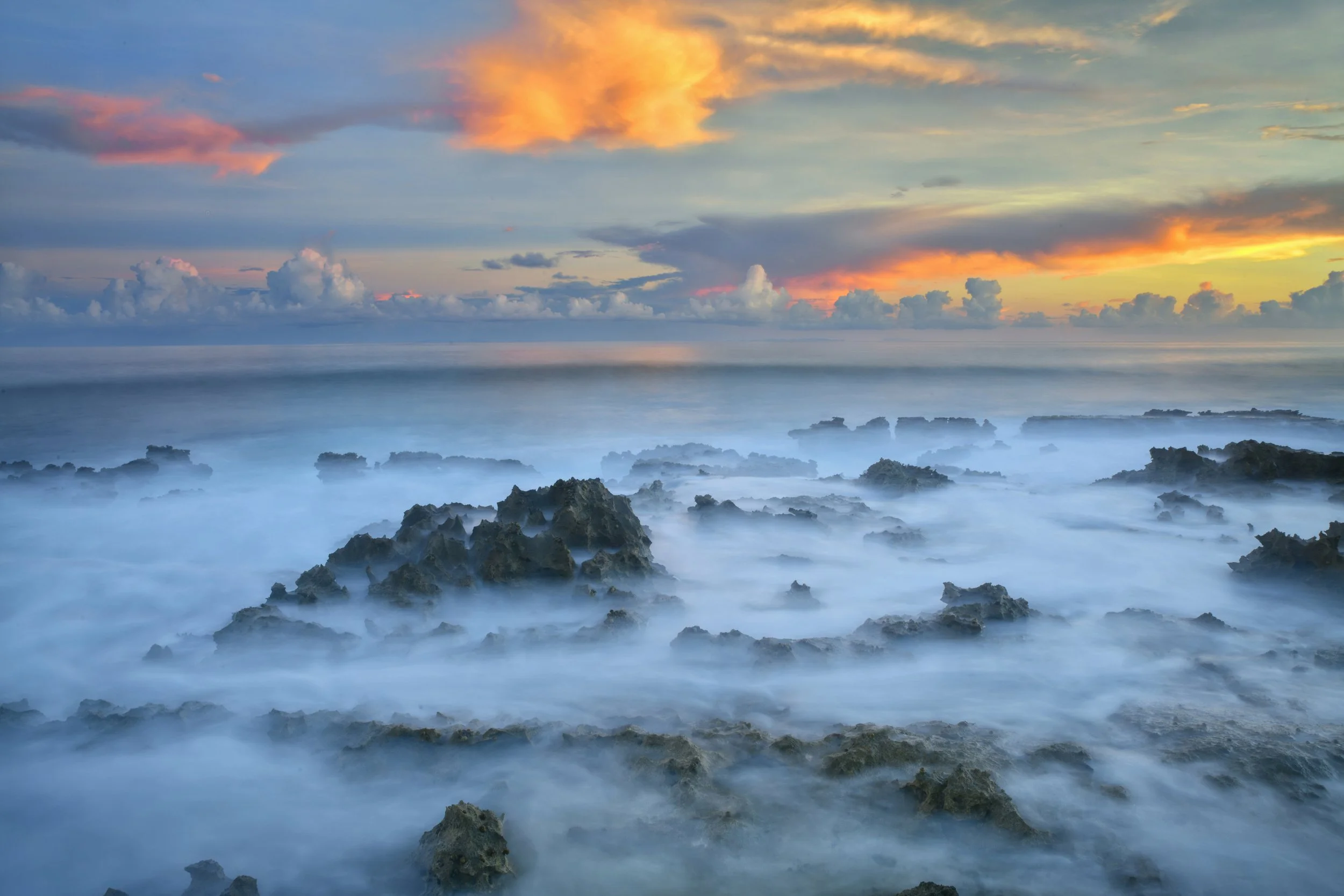 Sunset over a rocky shoreline with fog over the water and a colorful sky with clouds.