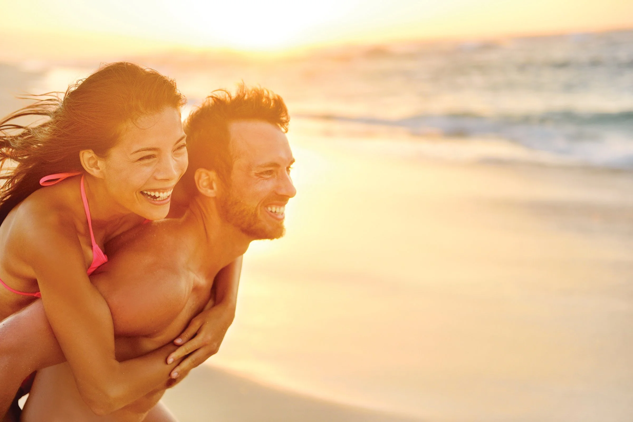 A smiling couple enjoying a piggyback ride on the beach during sunset, with the ocean in the background.