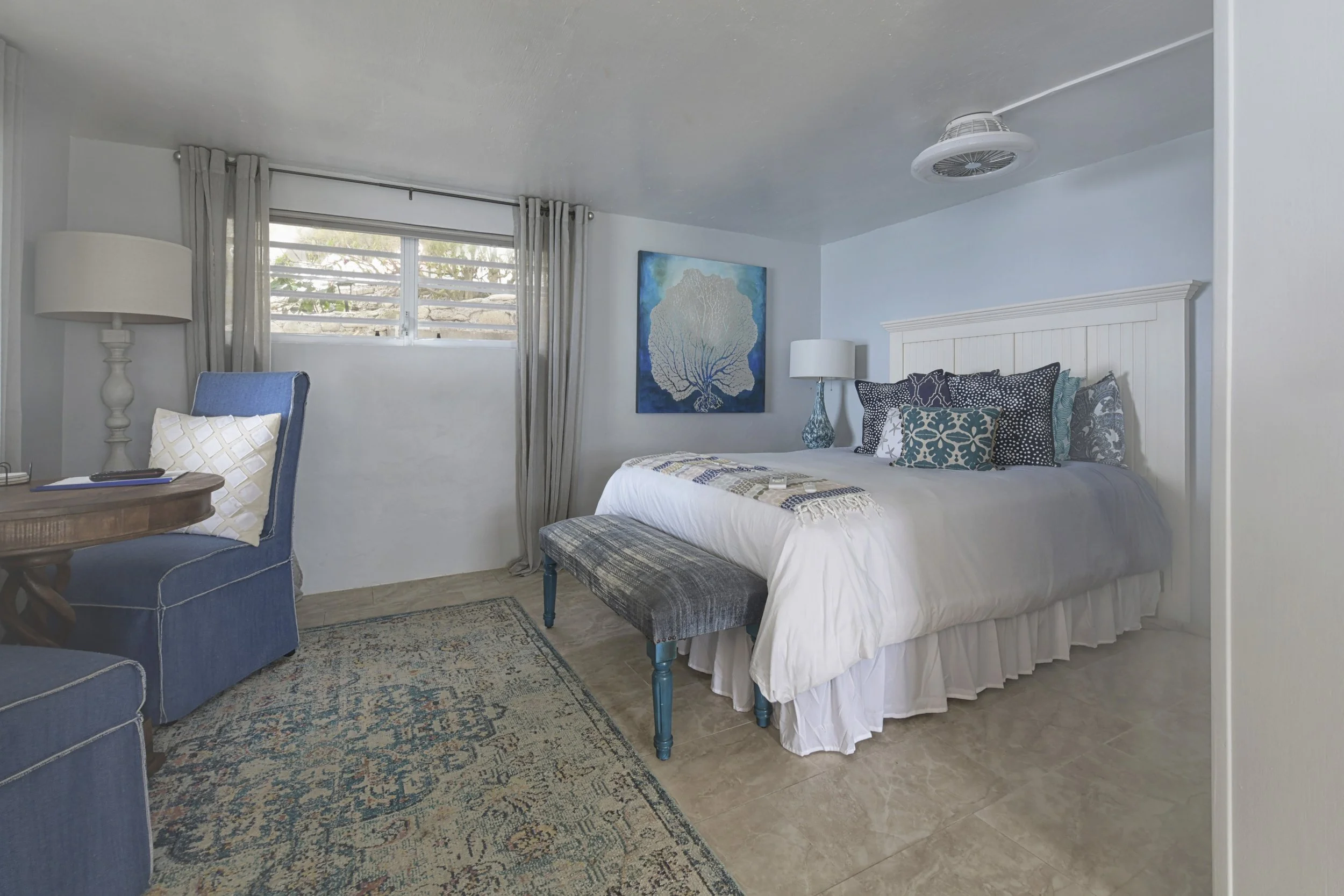 A bedroom with a white bed, blue pillows, a bedside lamp, a painting of a tree above the bed, a bench at the foot of the bed, a window with curtains, a blue armchair with a pillow, and a patterned rug on a tiled floor.