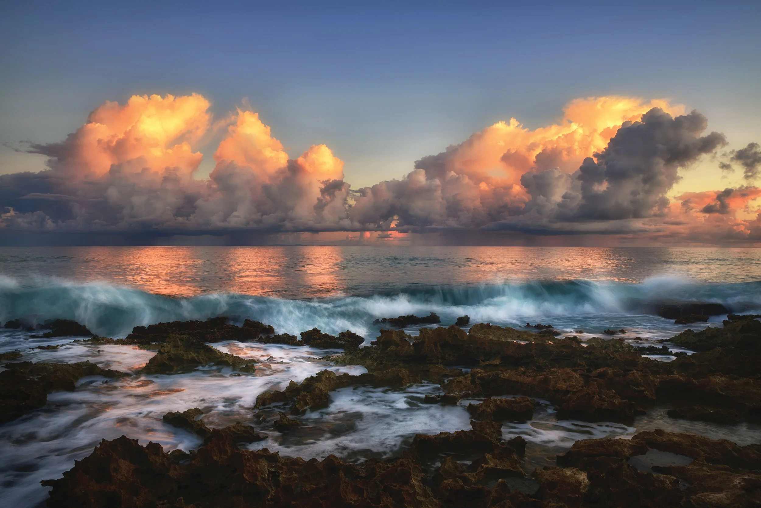 Sunset over the ocean with large clouds, waves crashing on rocky shore in the foreground.