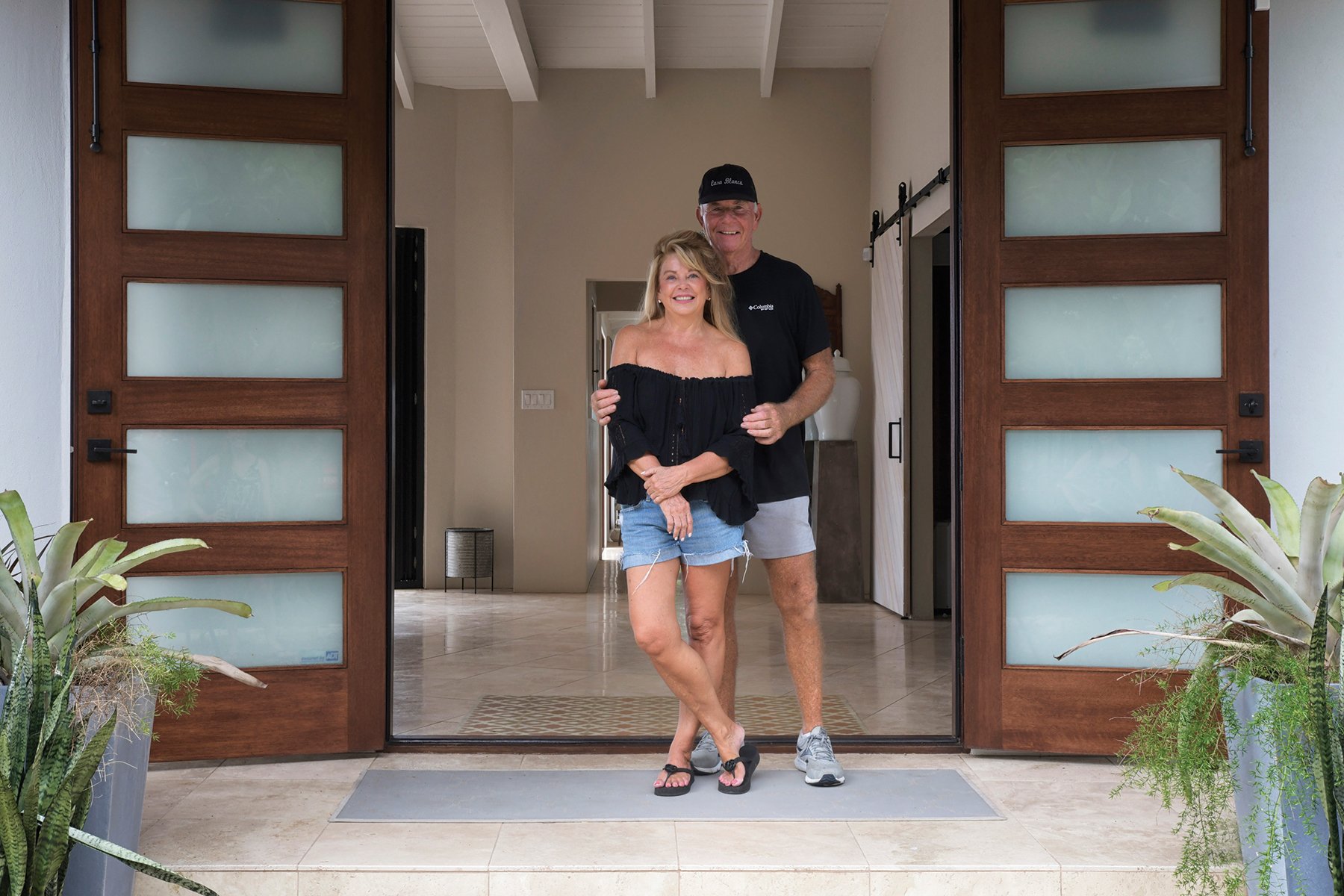 A smiling couple standing in the doorway of a modern house, surrounded by potted plants, with the man wearing a black cap and black T-shirt, and the woman in an off-the-shoulder black top and denim shorts.
