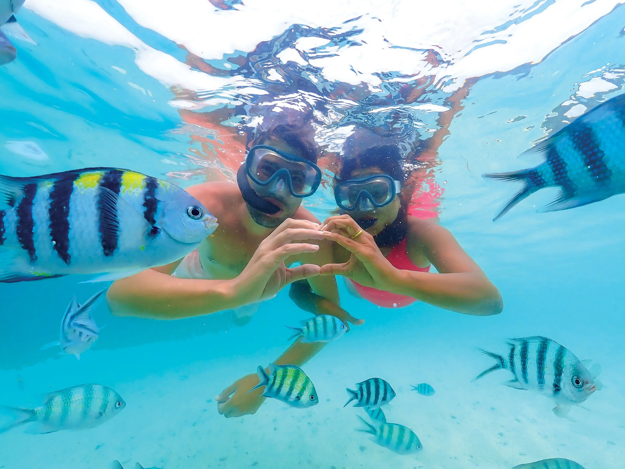 Two women snorkeling underwater among colorful fish, making a heart shape with their hands.
