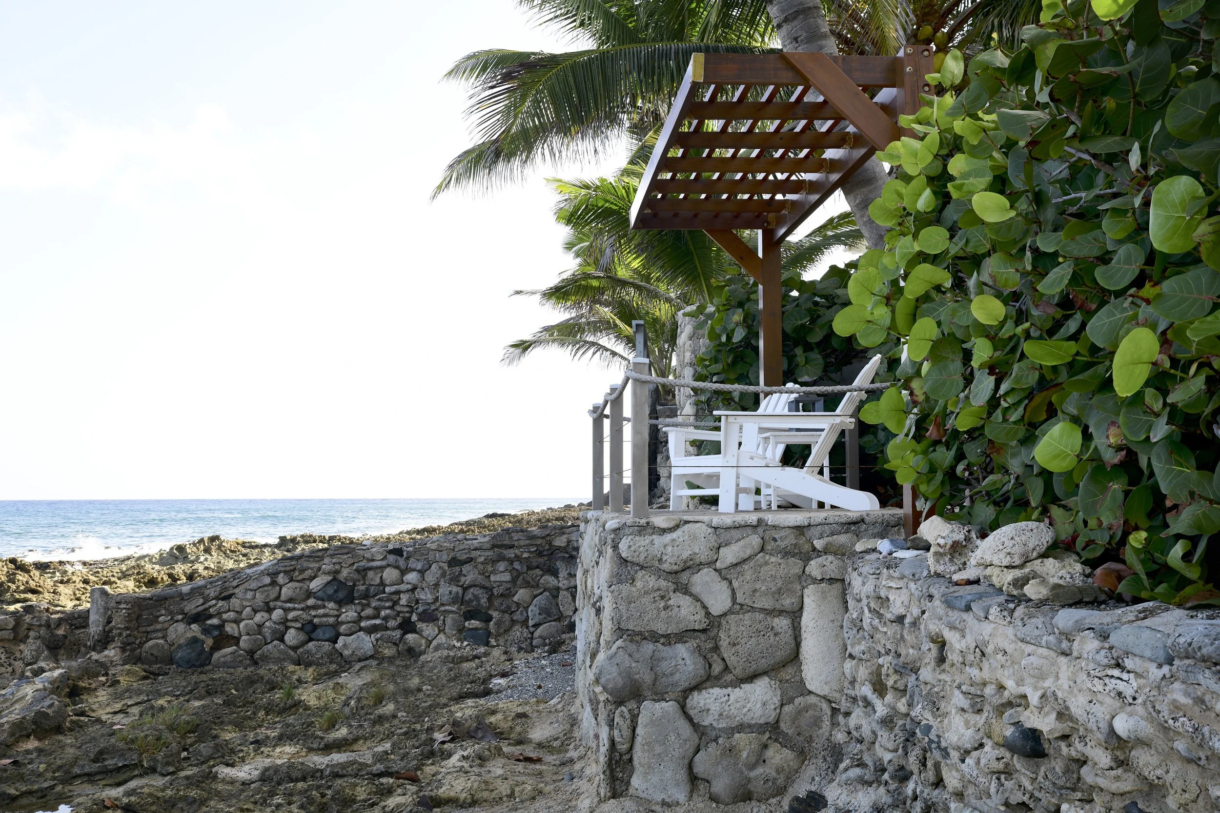 Coastal scene with a stone wall, white deck chairs, and a wooden pergola on a rocky shore, with green palm trees and dense foliage, overlooking the ocean.