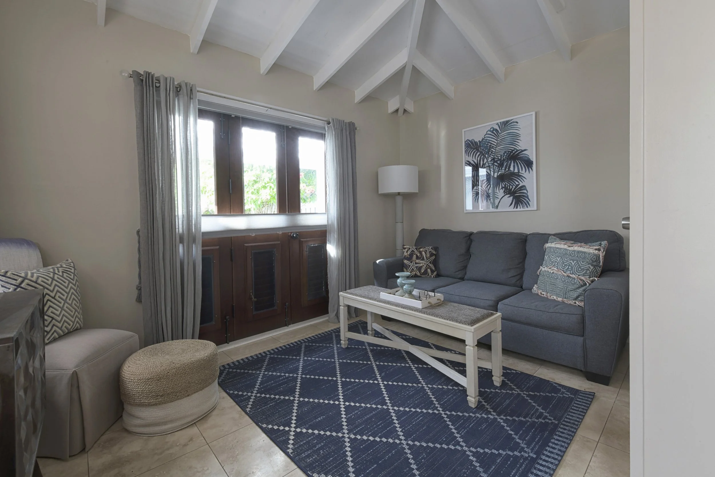 Living room with a blue sofa, white coffee table, patterned pillows, large window with curtains, framed tropical plant artwork, and a beige ottoman.