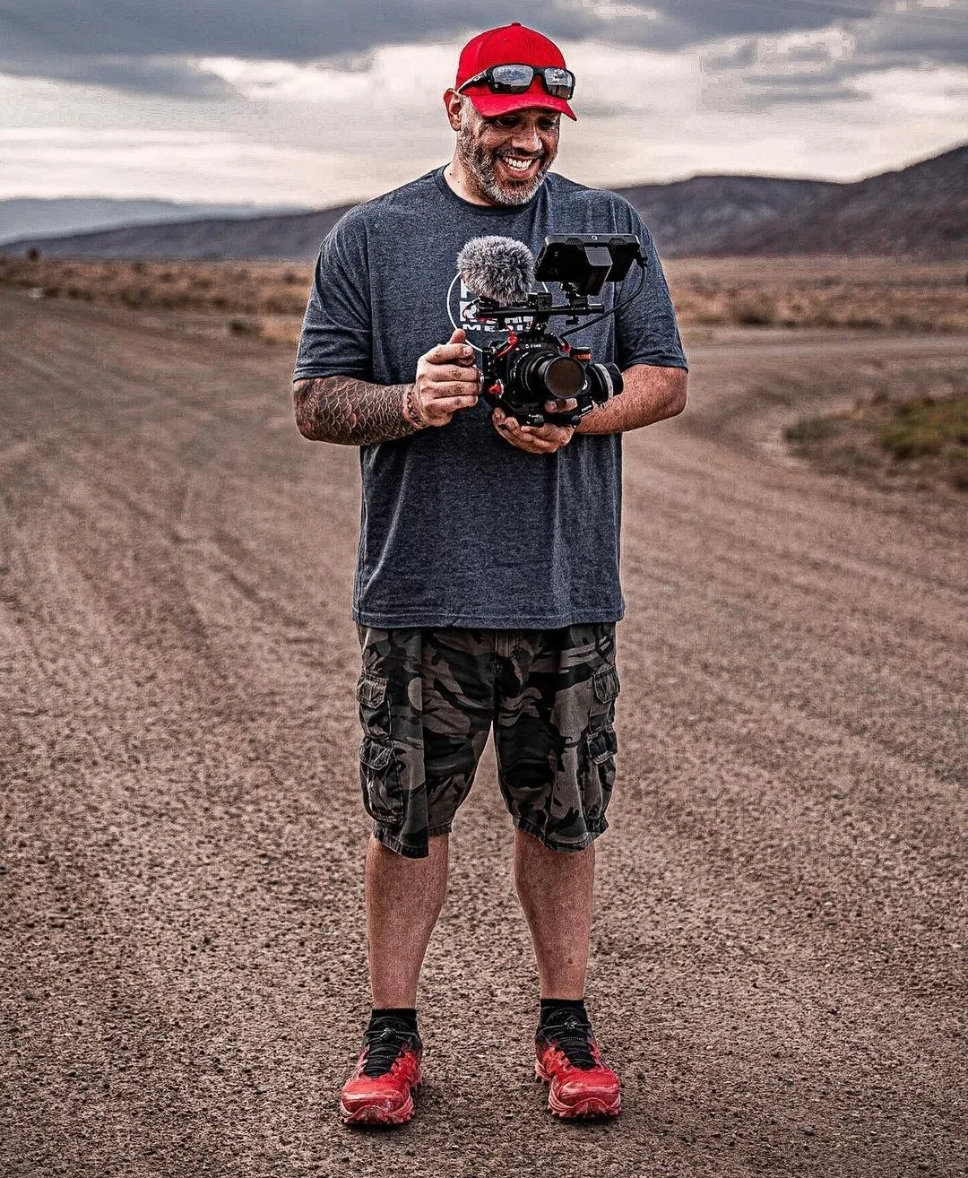 A man stands outdoors on a dirt road in a remote, mountainous area, holding a professional camera with a microphone and a monitor.