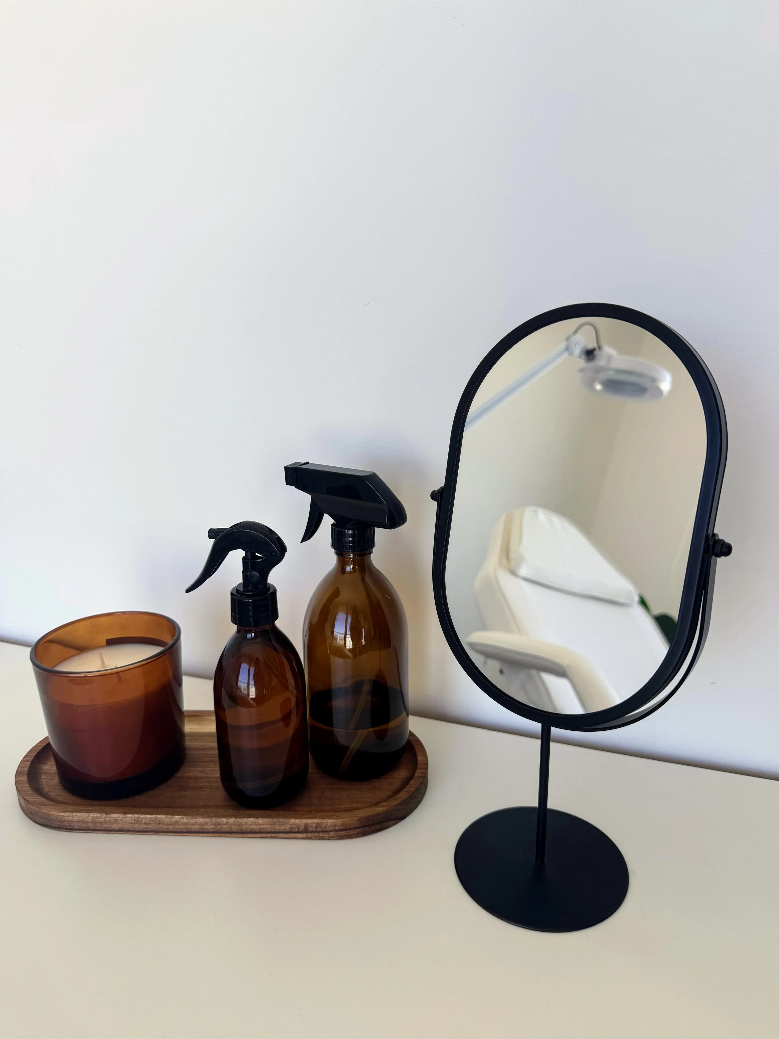 Espejo de escritorio, dos frascos de spray en tonos marrón, una vela en un recipiente de vidrio en tonos marrón y castaño, todos en una bandeja de madera sobre una mesa blanca, reflejados en un espejo de pie de negro, donde se puede ver una camilla.