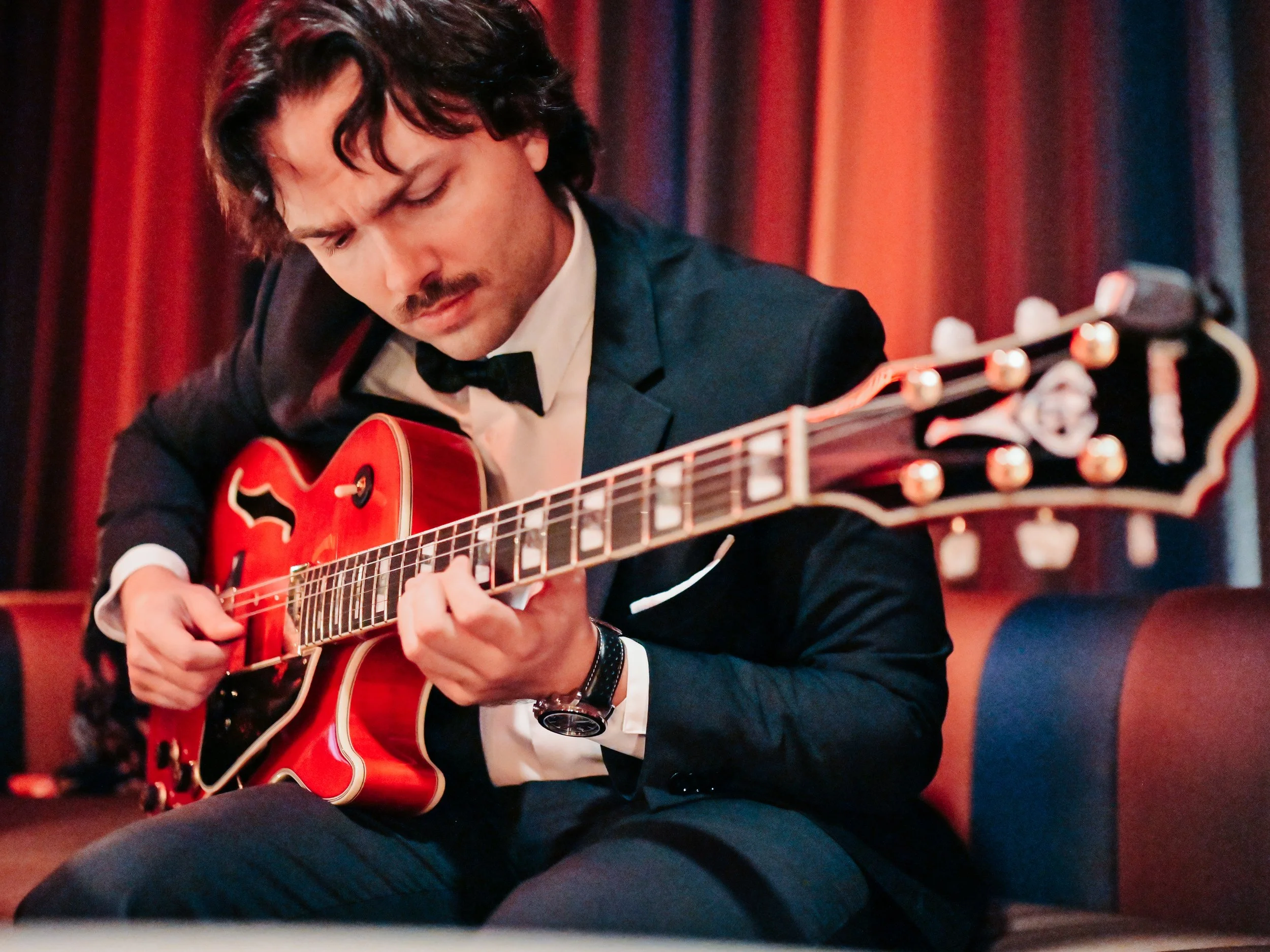Man in tuxedo playing a red hollow-body electric guitar in a dimly lit room with red curtains.