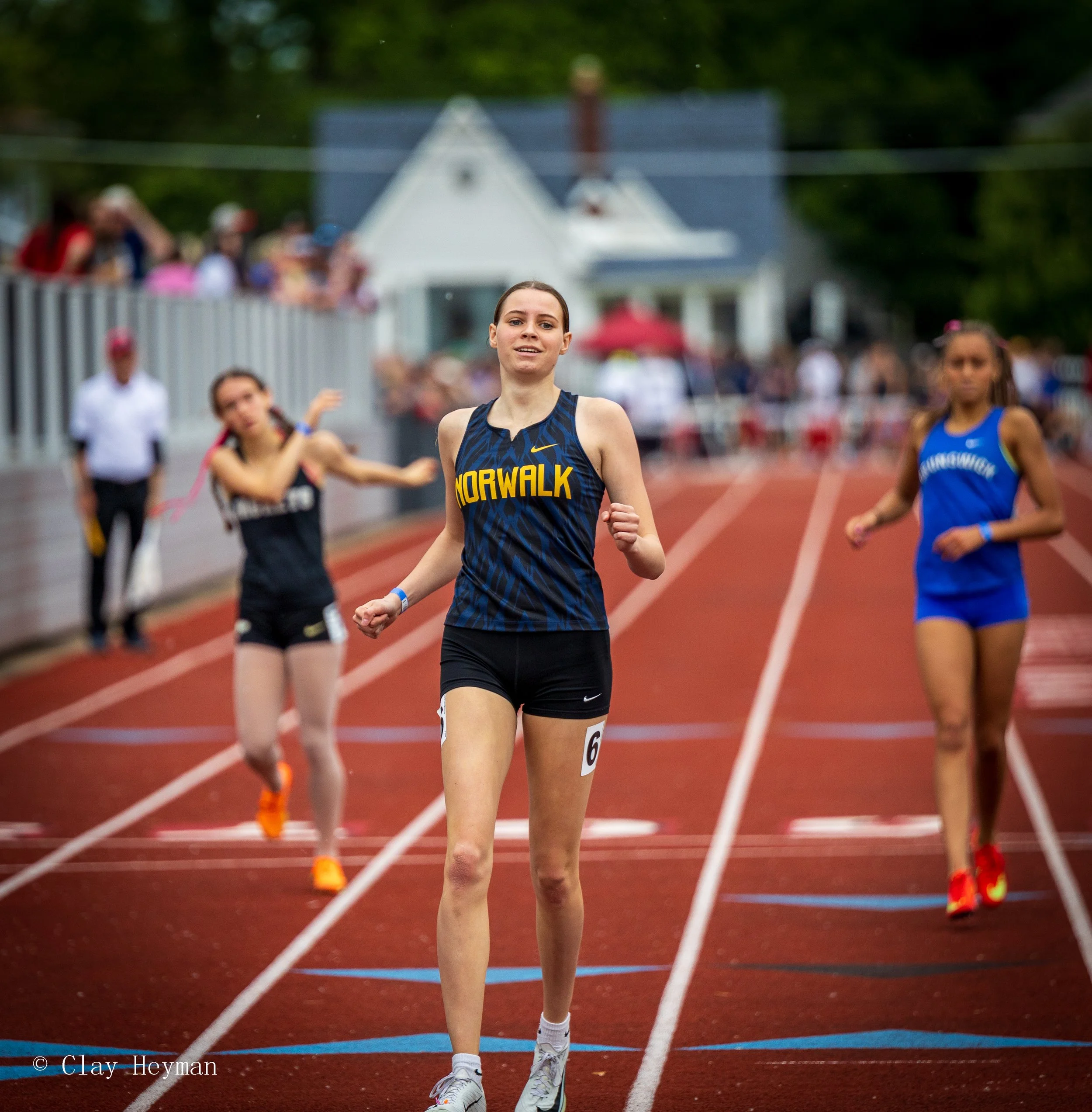 A young female athlete in a blue and yellow 'MorWalk' uniform running on a red track with other runners close behind during a race, with a crowd of spectators and a house in the background.