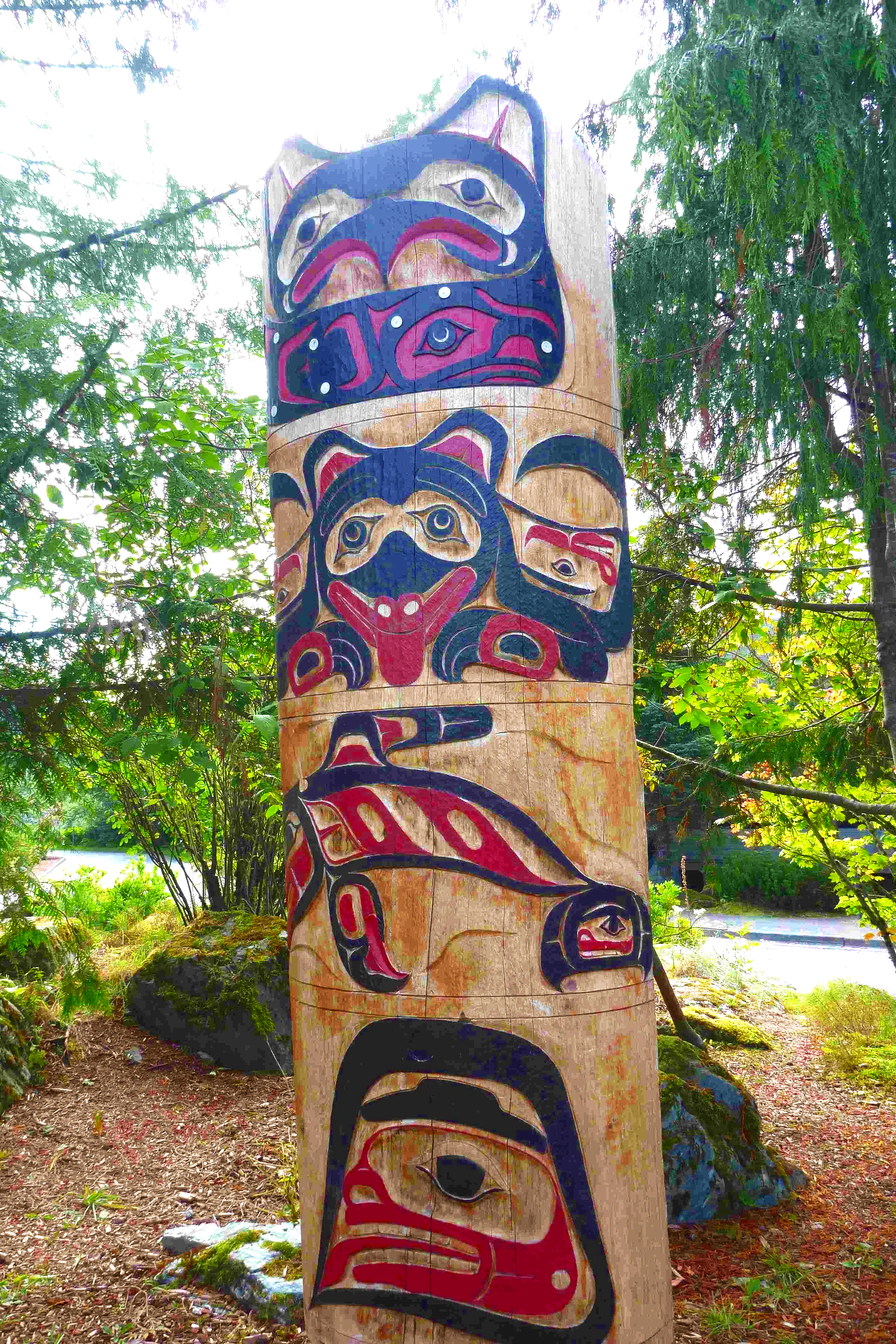 Colorful indigenous totem pole standing outdoors among green trees and bushes. Alaska