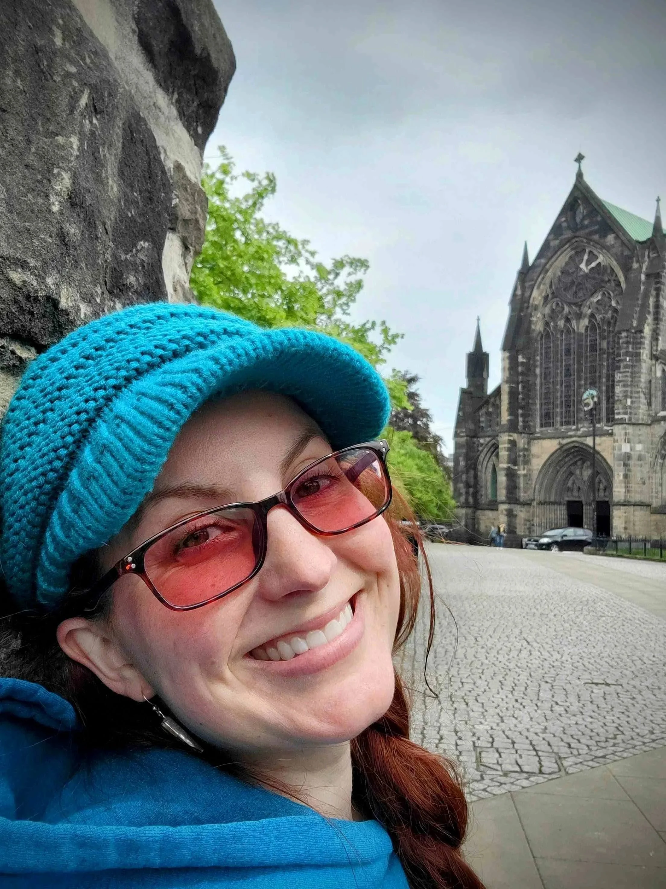 A woman with red hair, wearing rose colored glasses and a bright blue hoodie, smiling in front of the gothic Glasgow Cathedral a historic church, situated on cobblestone Castle Street in Glasgow Scotland.
