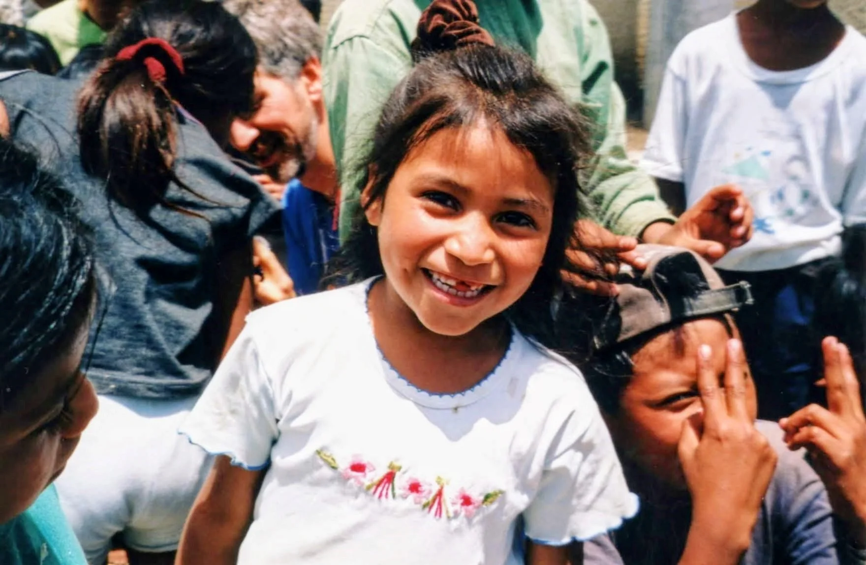 Smiling young girl with curly hair and a white shirt surrounded by other children in a crowd.