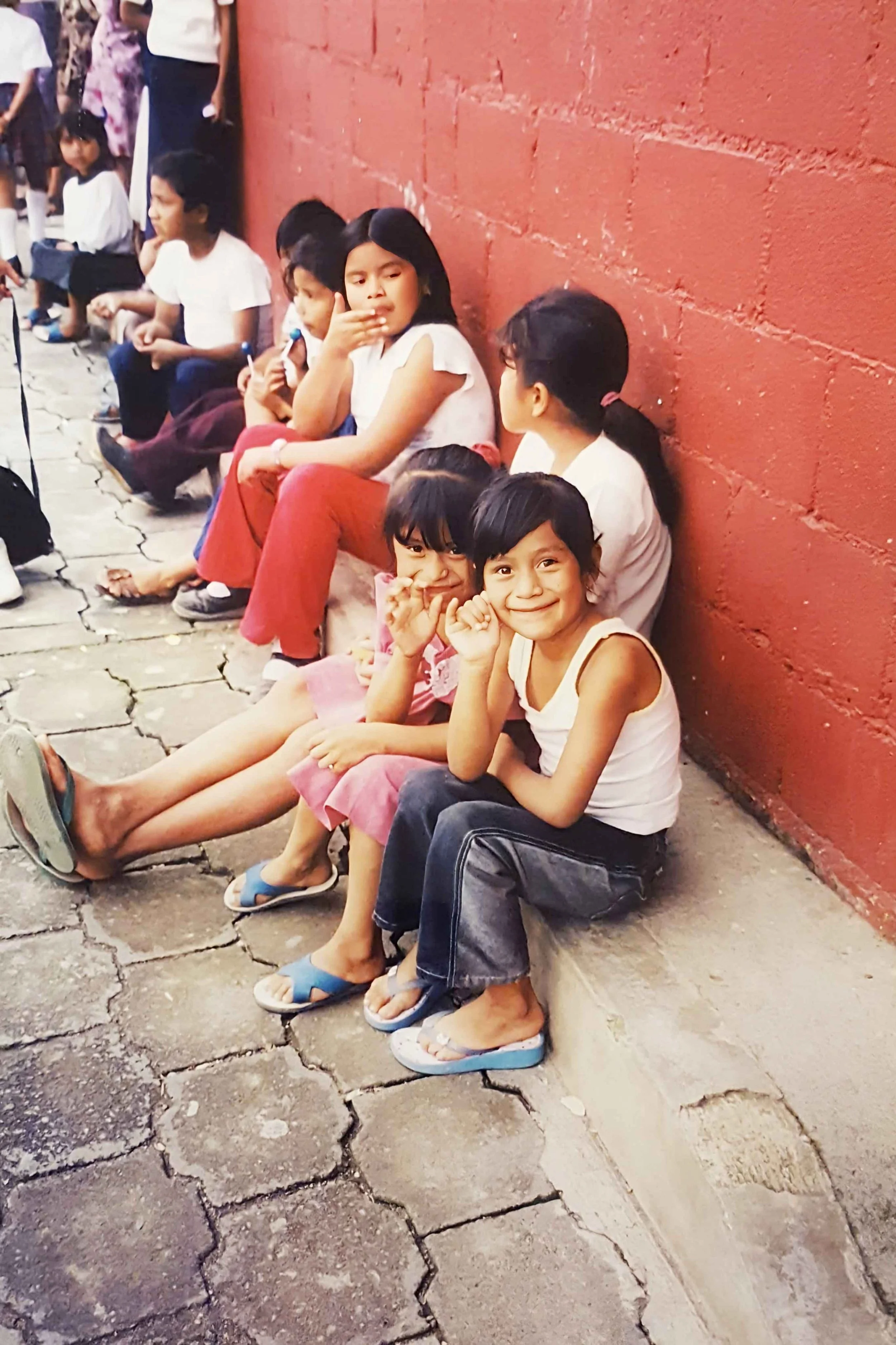 Children sitting on a sidewalk against a red brick wall, some are smiling and making gestures, with varied appearances and casual clothing.