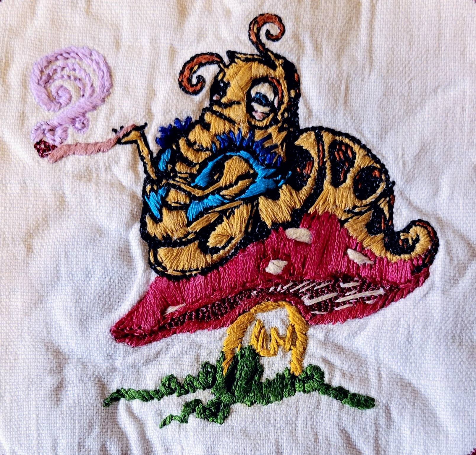 Embroidery of a caterpillar sitting on a red mushroom with white spots, wearing a blue top, yellow skin, and holding a purple spiral in his hand, on a white fabric background.