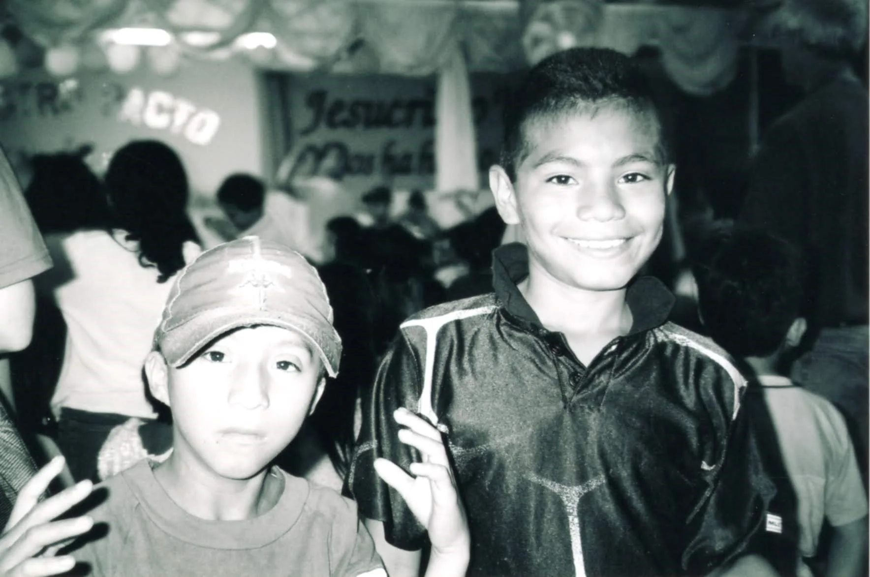 Two boys smiling and making peace signs at a crowded community event or festival. One boy is wearing a baseball cap and a t-shirt, the other is wearing a shiny windbreaker. The background has a banner with partially visible text and other people in attendance.
