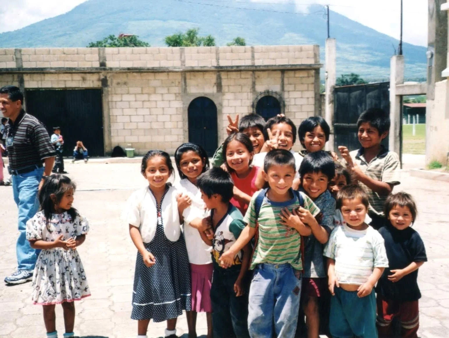 Group of smiling children and a man standing outdoors in front of a brick building with hills in the background