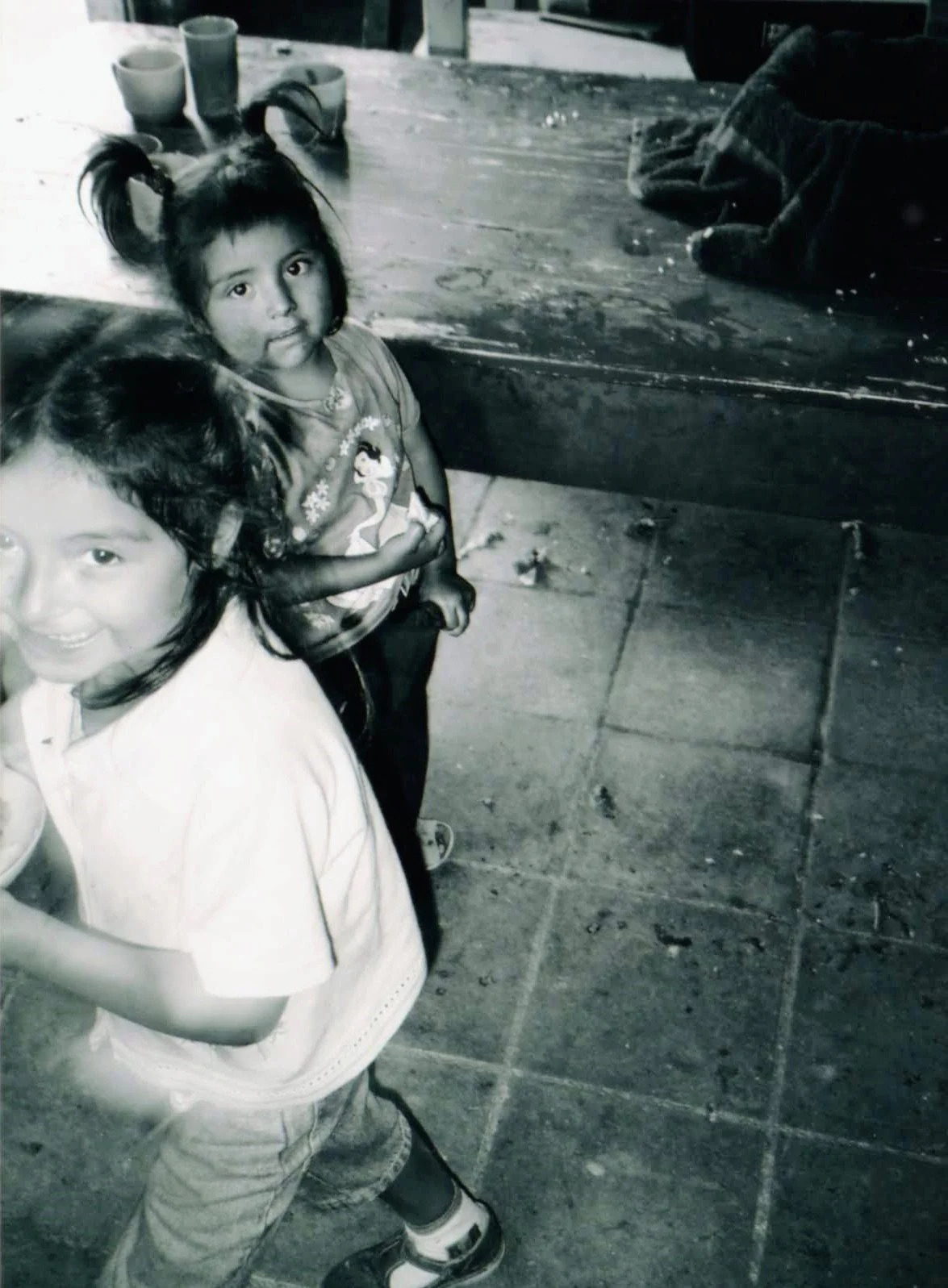 Two young girls standing outdoors on a sidewalk near a wooden table with cups on it. Both are smiling and looking at the camera.