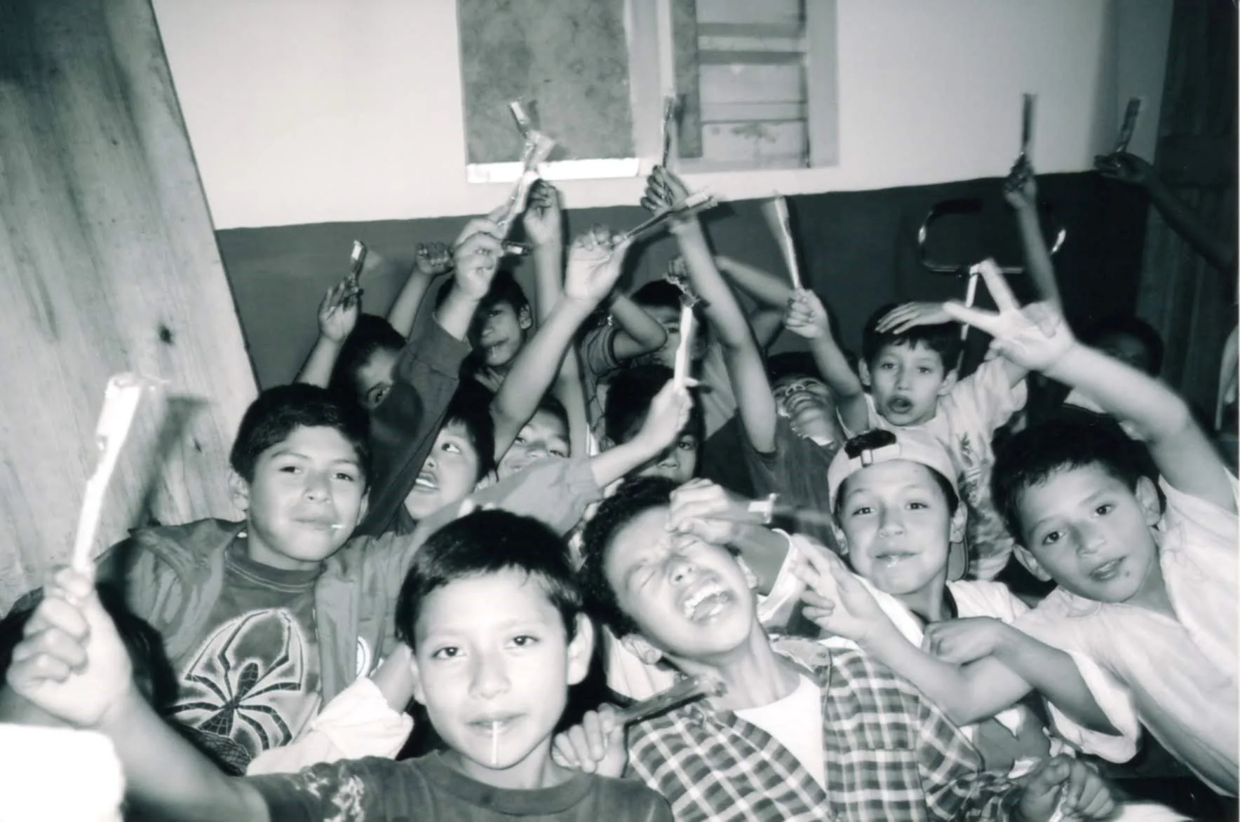 A group of children gathered closely together, smiling and holding toothbrushes, with some making peace signs, in an indoor setting.