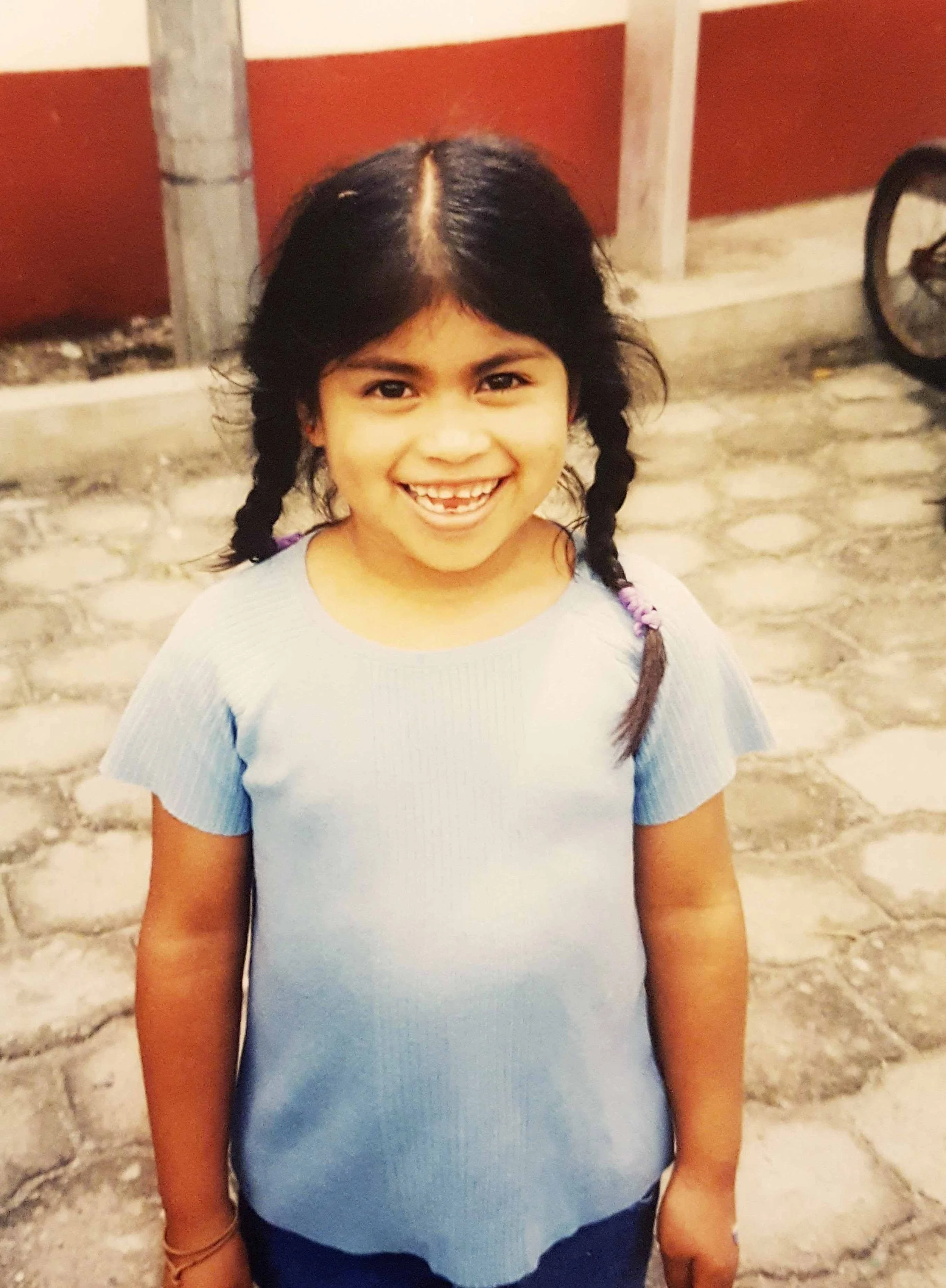 A young girl with dark hair in two braids smiling outdoors in front of a stone wall and a red and white building.