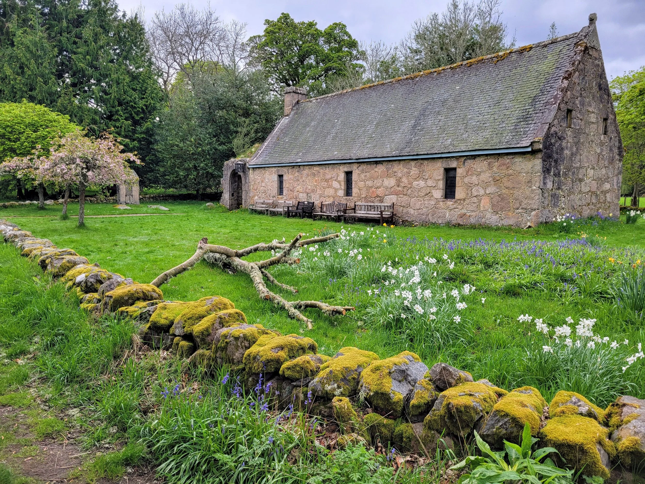 A stone church with a steeply pitched roof situated in a lush garden. The garden features a small, blossoming tree, colorful flowers, and a moss-covered stone wall. There are multiple benches along the side of the church, and tall trees surround the scene.