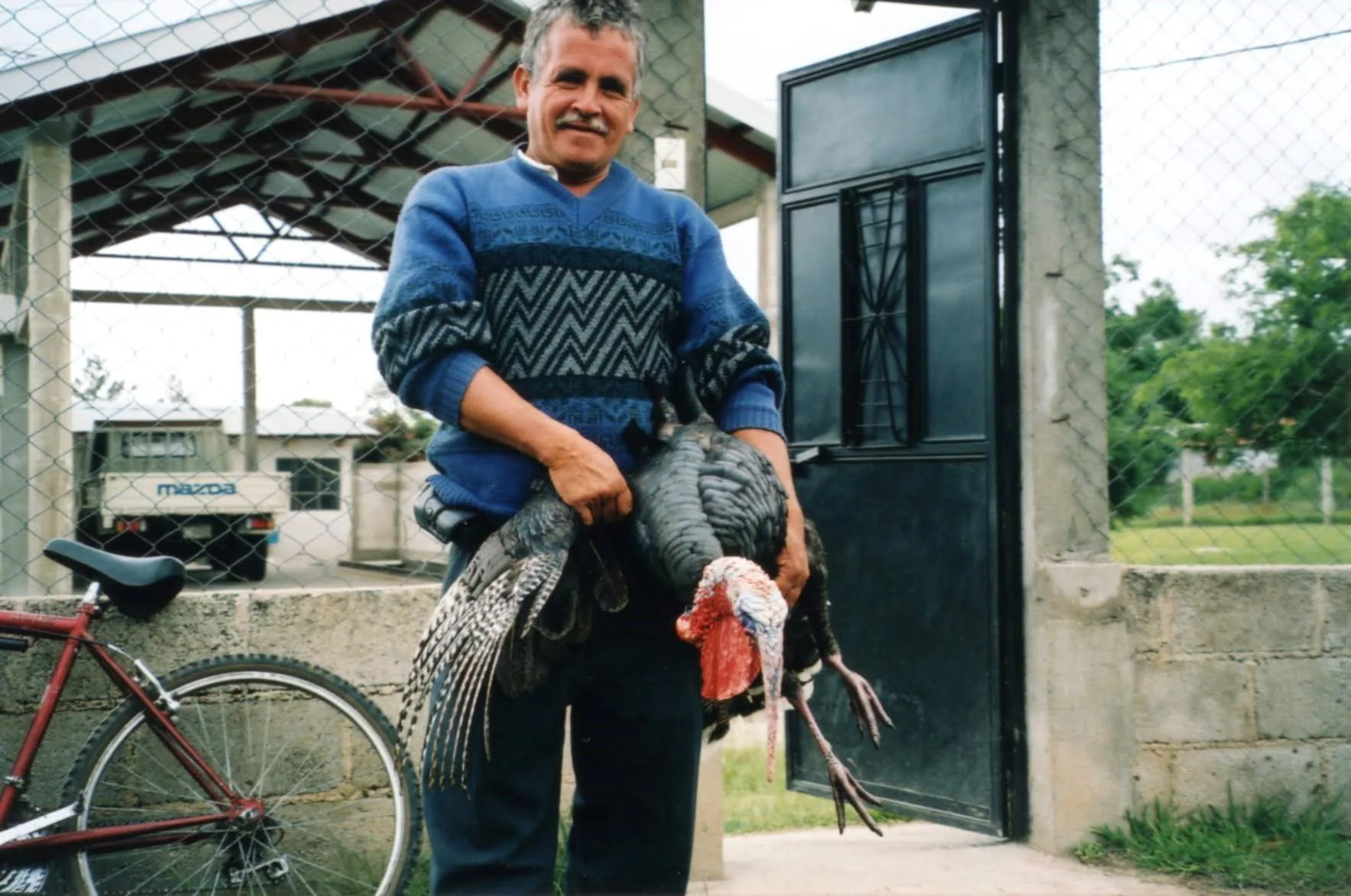 A man holding a turkey outside a gated area with a black door, a bicycle, and a white Mazda truck in the background.