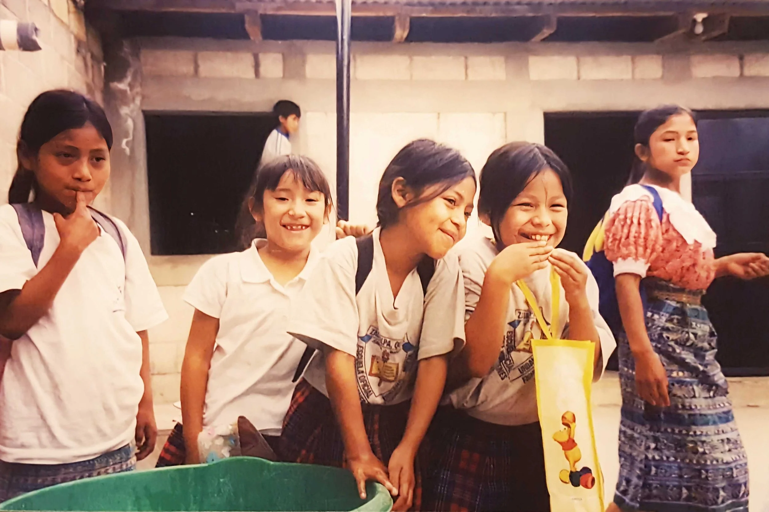 A group of five girls standing side by side indoors, some smiling and some with neutral or slightly serious expressions, with a boy in the background. They are dressed casually and appear to be at a school or community event.